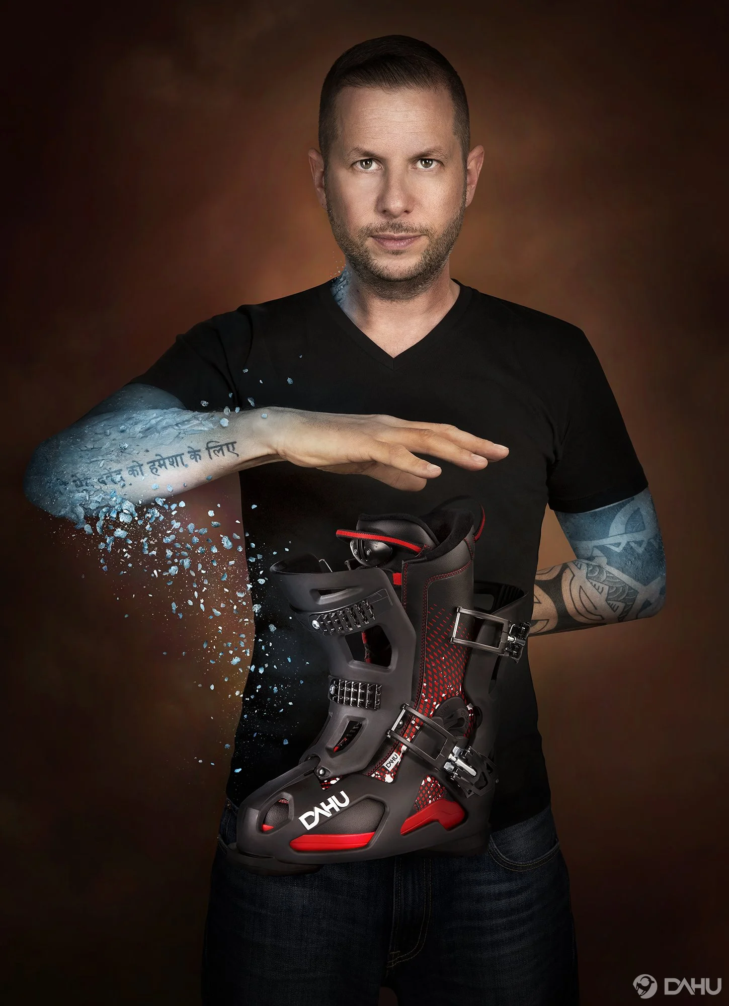 Un homme avec un tatouage sur l'avant-bras pose avec une chaussure de ski sur le ventre, portant un VTT et portant une expression confiante. Il a une barbe légère et est vêtu d'un t-shirt noir. La photo a un fond sombre avec un dégradé chaud.
