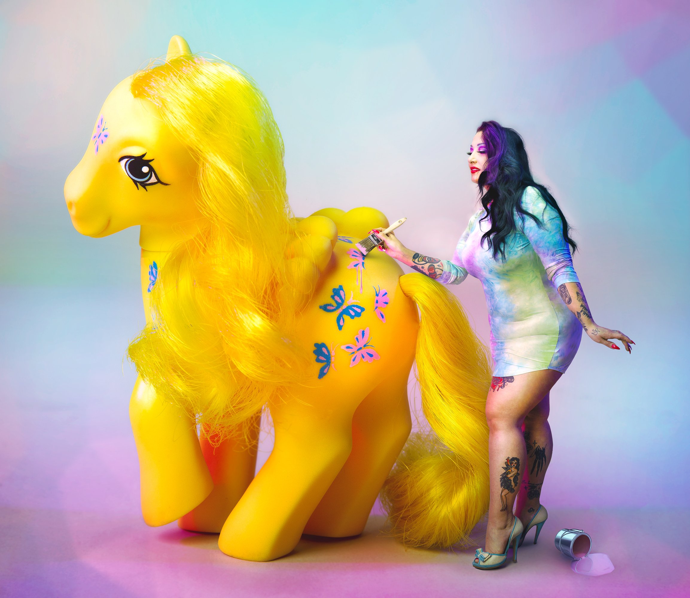 Une femme avec des tatouages colorés, cheveux violets et noirs, en train de peindre un bonhomme de bonheur gonflable jaune avec une peinture blanche et un pinceau. La femme porte une robe tie-dye avec des talons bleus et des accessoires.