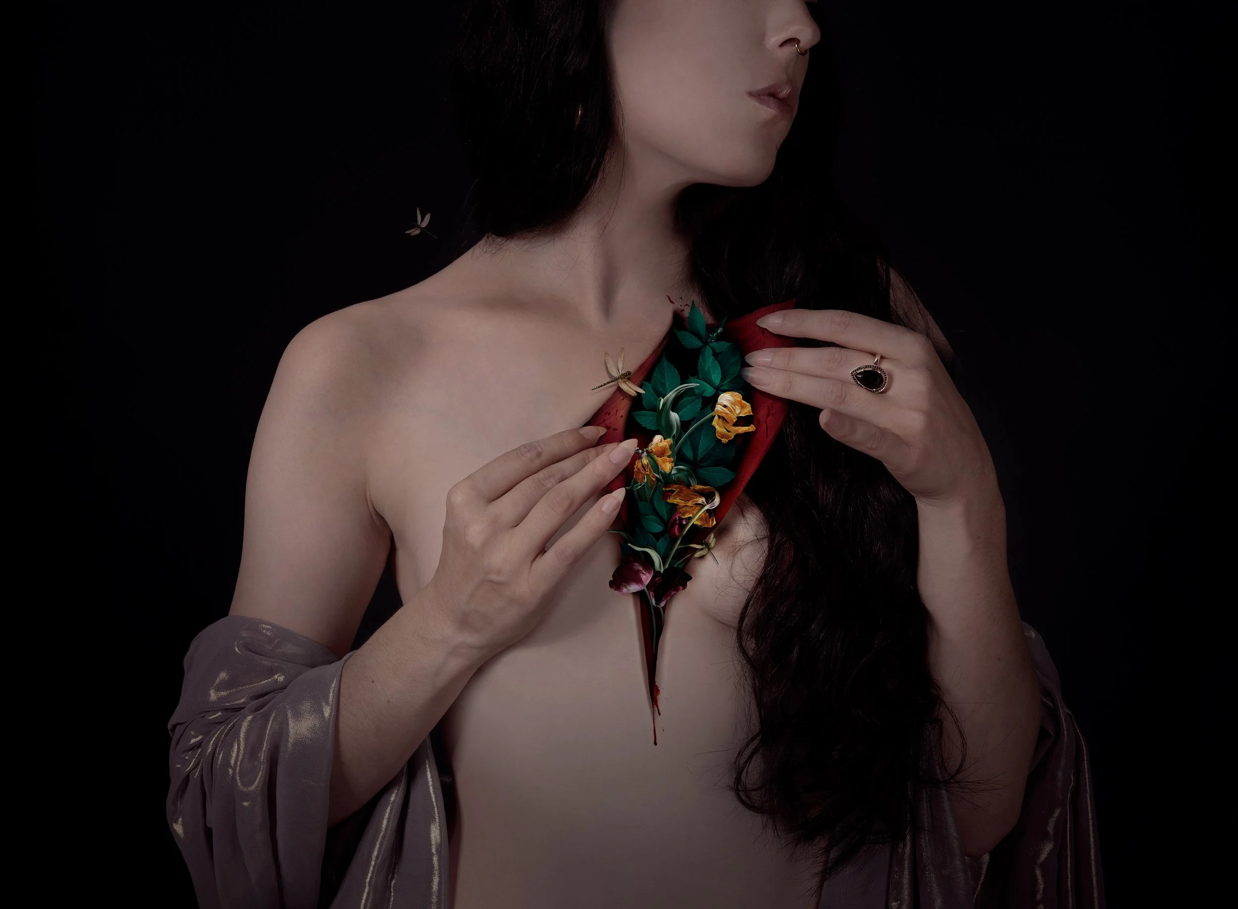 Une femme avec des cheveux longs et foncés, portant une robe à manches longues en satin gris, tenant ses mains sur sa poitrine. La photo est centrée sur le haut du corps et le visage est partiellement visible, avec un fond noir.