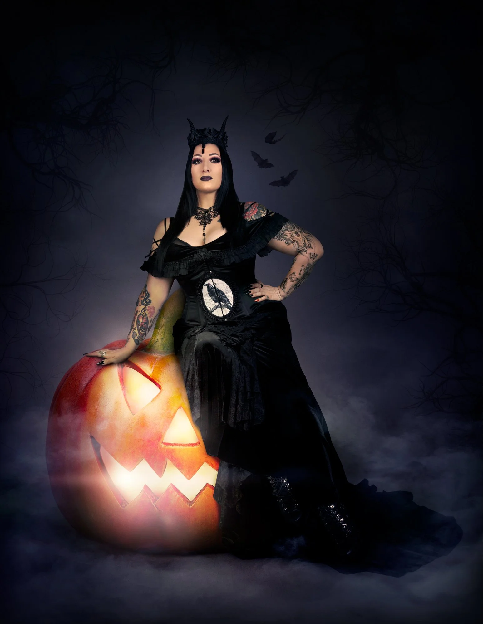 Une femme gothique avec un maquillage sombre et des tatouages, portant une couronne en forme de cornes et une robe noire, pose devant une grande citrouille sculptée avec un visage de Halloween, dans un décor nocturne sombre avec des branches d'arbres