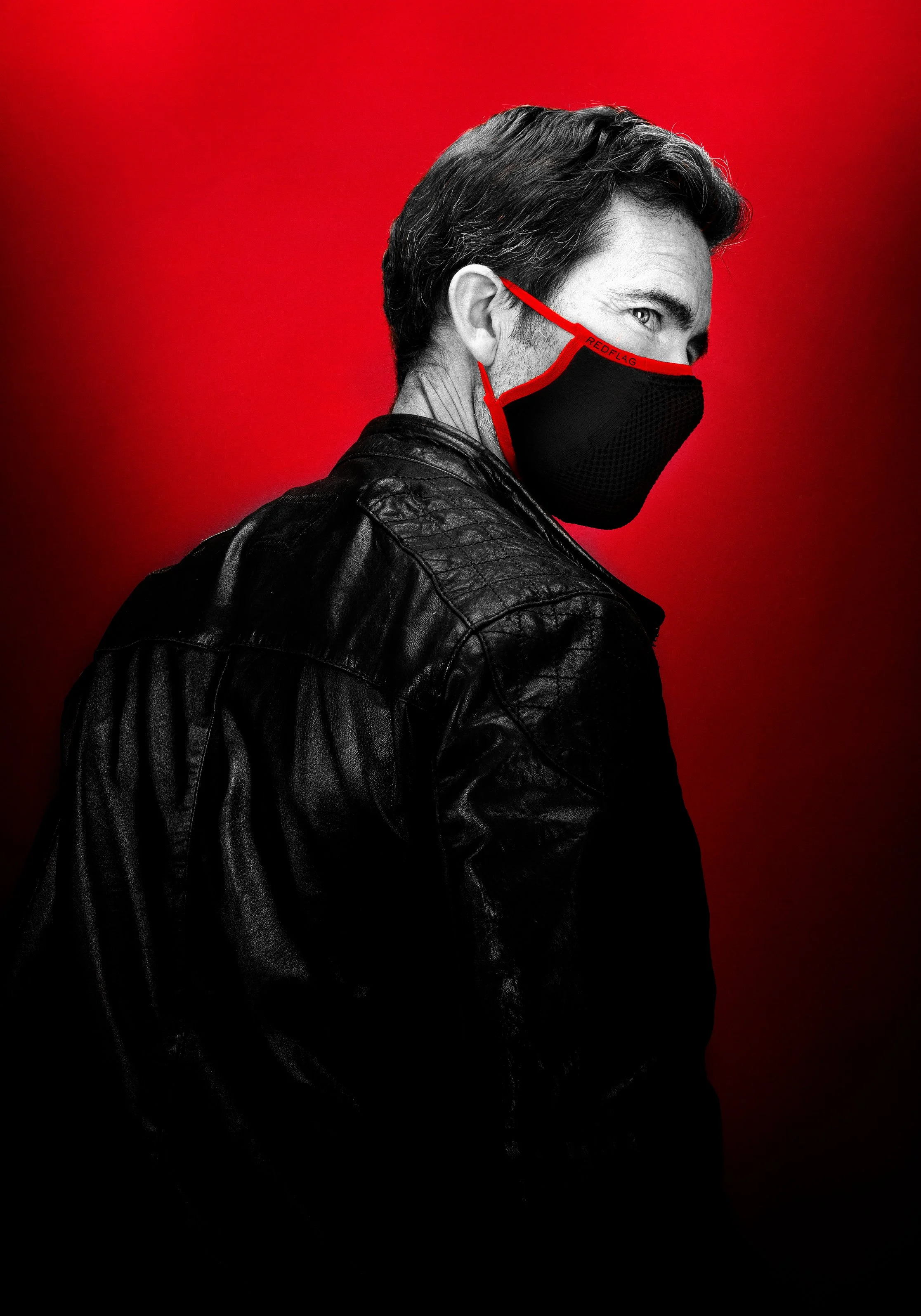 Portrait d'un homme portant un masque noir avec une bordure rouge, vêtement en cuir noir, fond rouge.