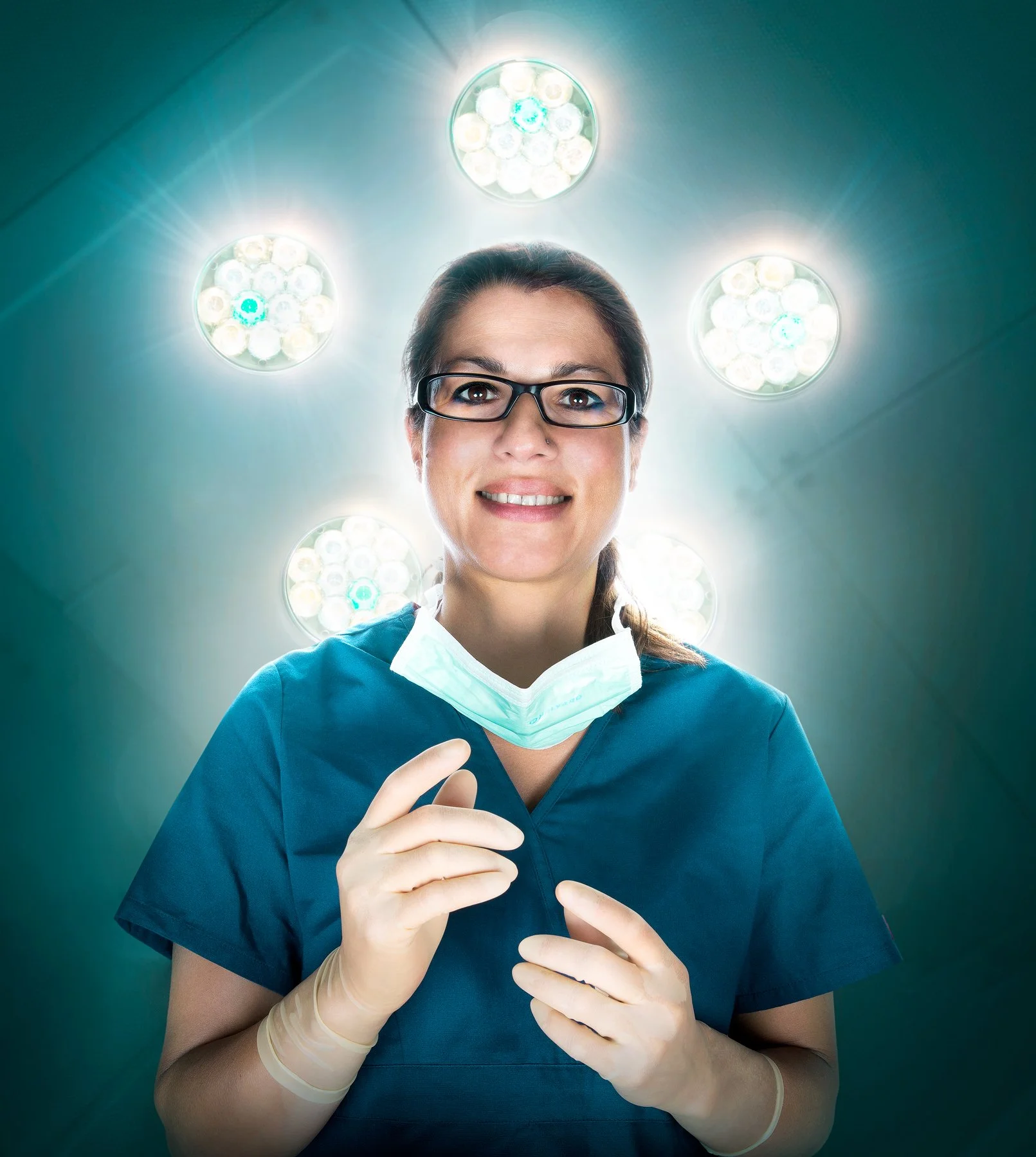 Une femme médecin souriante portant des gants chirurgicaux, une blouse bleue, une paire de lunettes, et un masque facial abaissé. Elle se trouve dans une salle d'opération, avec des lampes chirurgicales au plafond