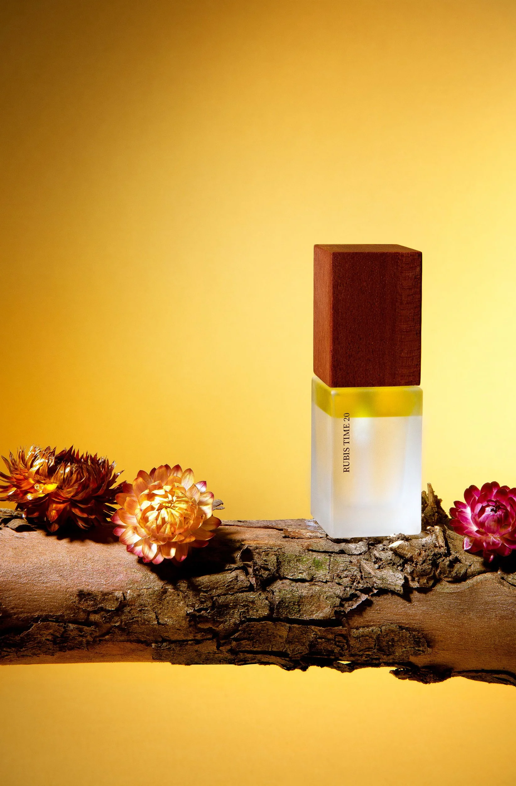 Un flacon de parfum blanc avec un bouchon en bois sur un morceau de bois, entouré de fleurs colorées, avec un fond jaune