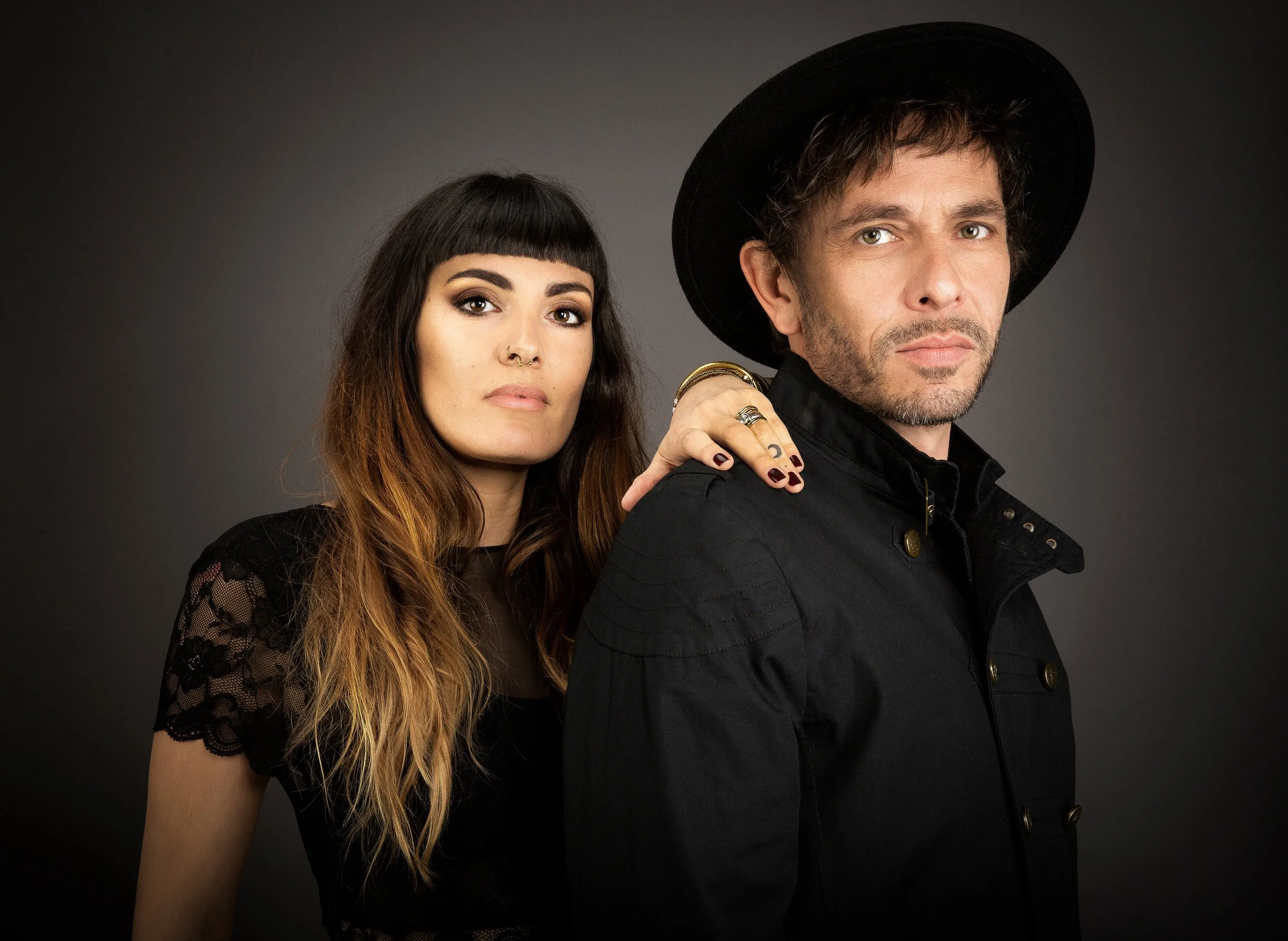 Un homme et une femme posant ensemble contre un fond gris sombre, l'homme porte un chapeau noir et une veste noire, et la femme a des cheveux longs et ondulés avec une coupe courte sur le devant.