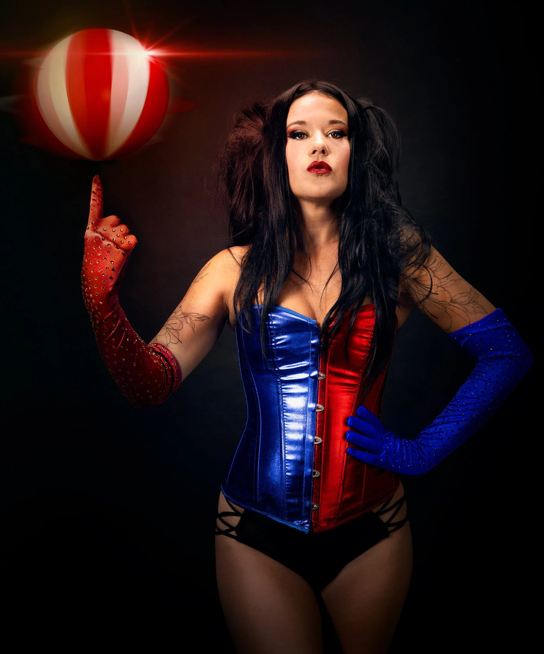 Femme avec tatouages en corset rouge et bleu, gants rouges et bleus, pointant vers une balle de baseball à rayures rouges et blanches, sur fond noir.