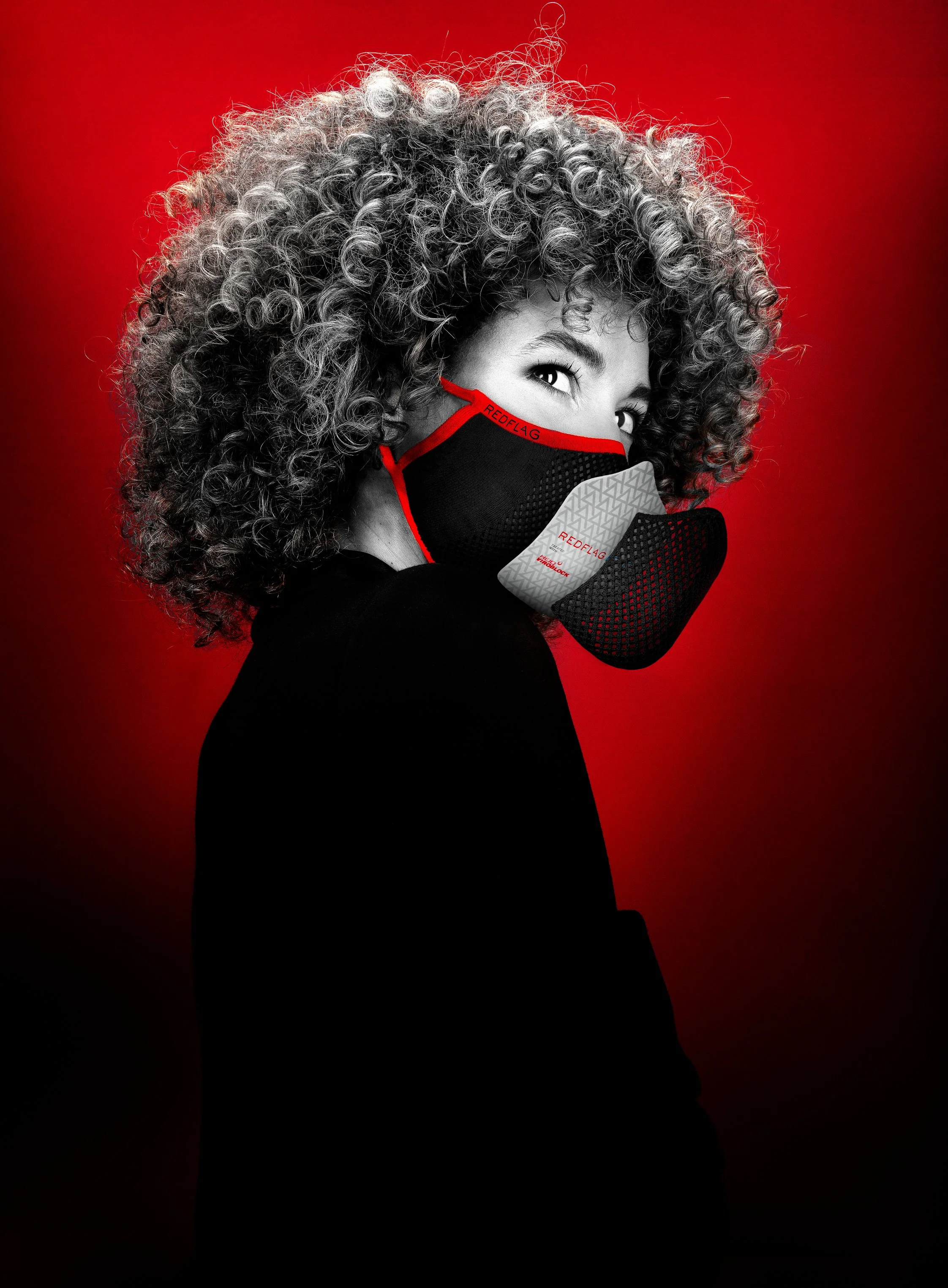 Une femme à cheveux bouclés porte un masque facial noir avec des détails rouges et une écharpe. Elle regarde par-dessus son épaule, avec un arrière-plan rouge profond.