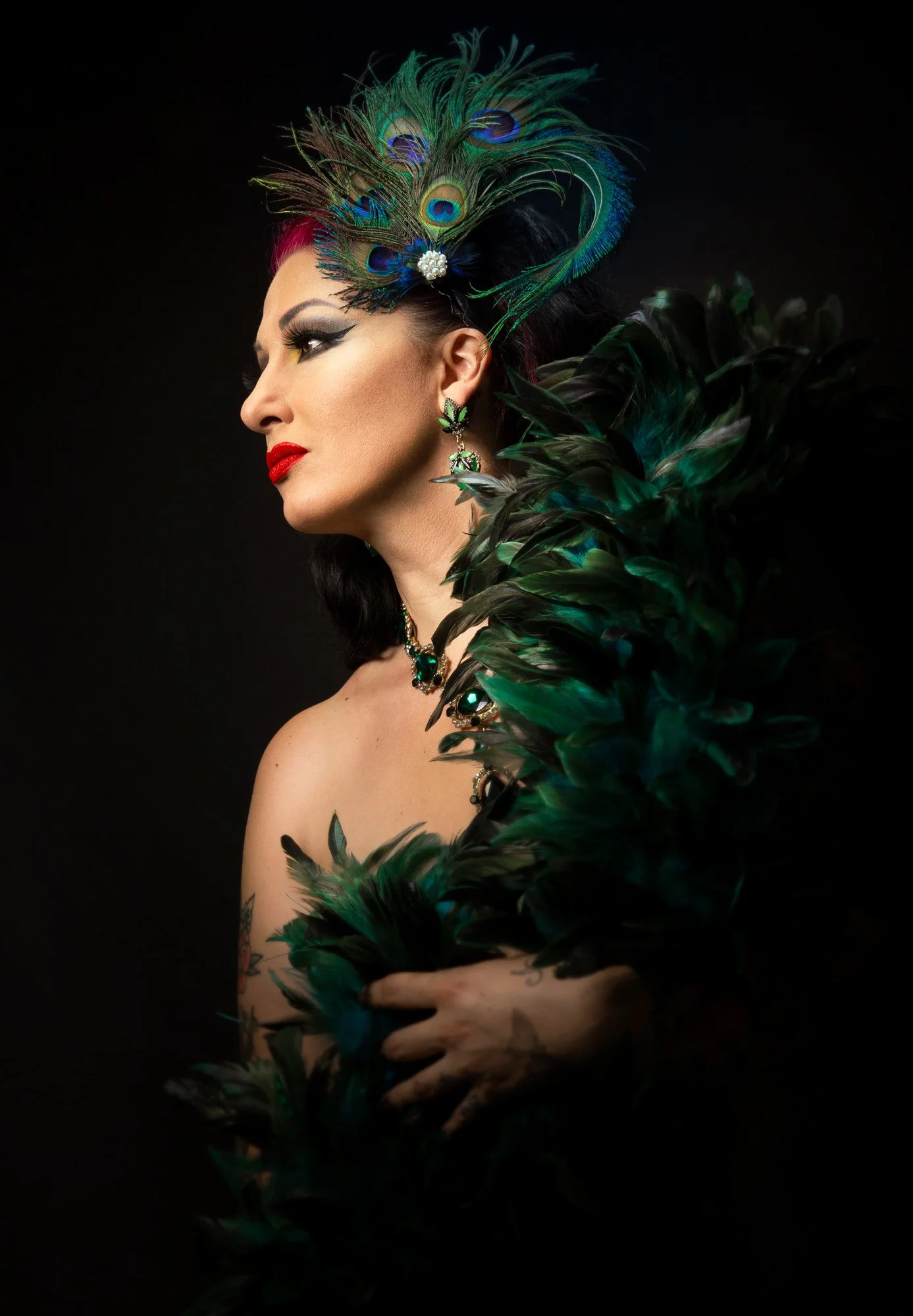 Portrait d'une femme portant un accessoire de cheveux en plumes de paon, des bijoux et entourée de plumes vertes, avec un maquillage marqué et un fond noir.