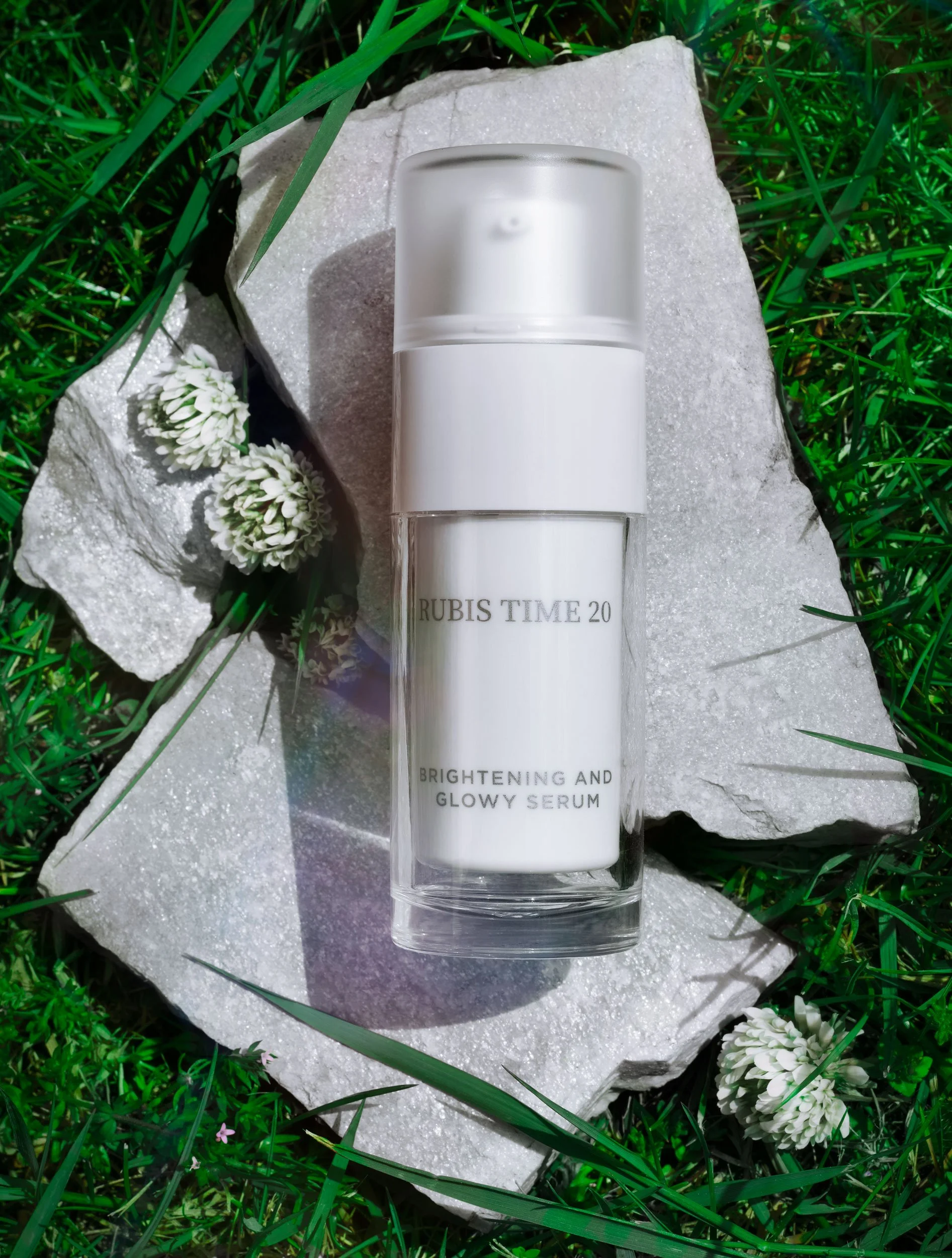 Un flacon de sérum blanc avec un capuchon en plastique, posé sur une pierre blanche, entouré d'herbe verte et de petites fleurs blanches.
