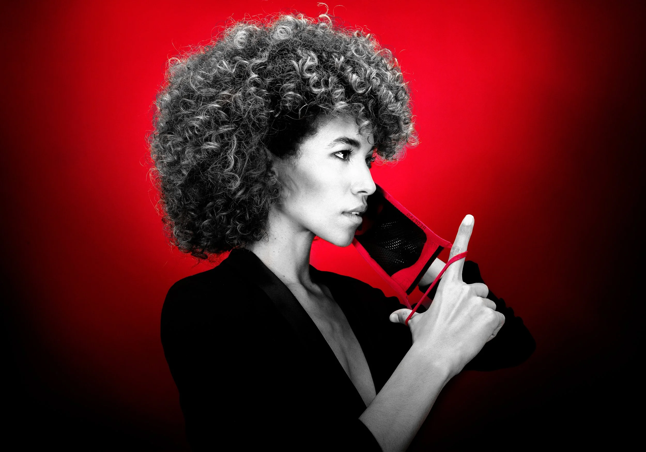 Une femme aux cheveux bouclés gris foncé, portant une veste noire, tient un fil rouge et semble faire un geste de silence avec son index. Le fond est rouge et noir, avec une effet de dégradé.