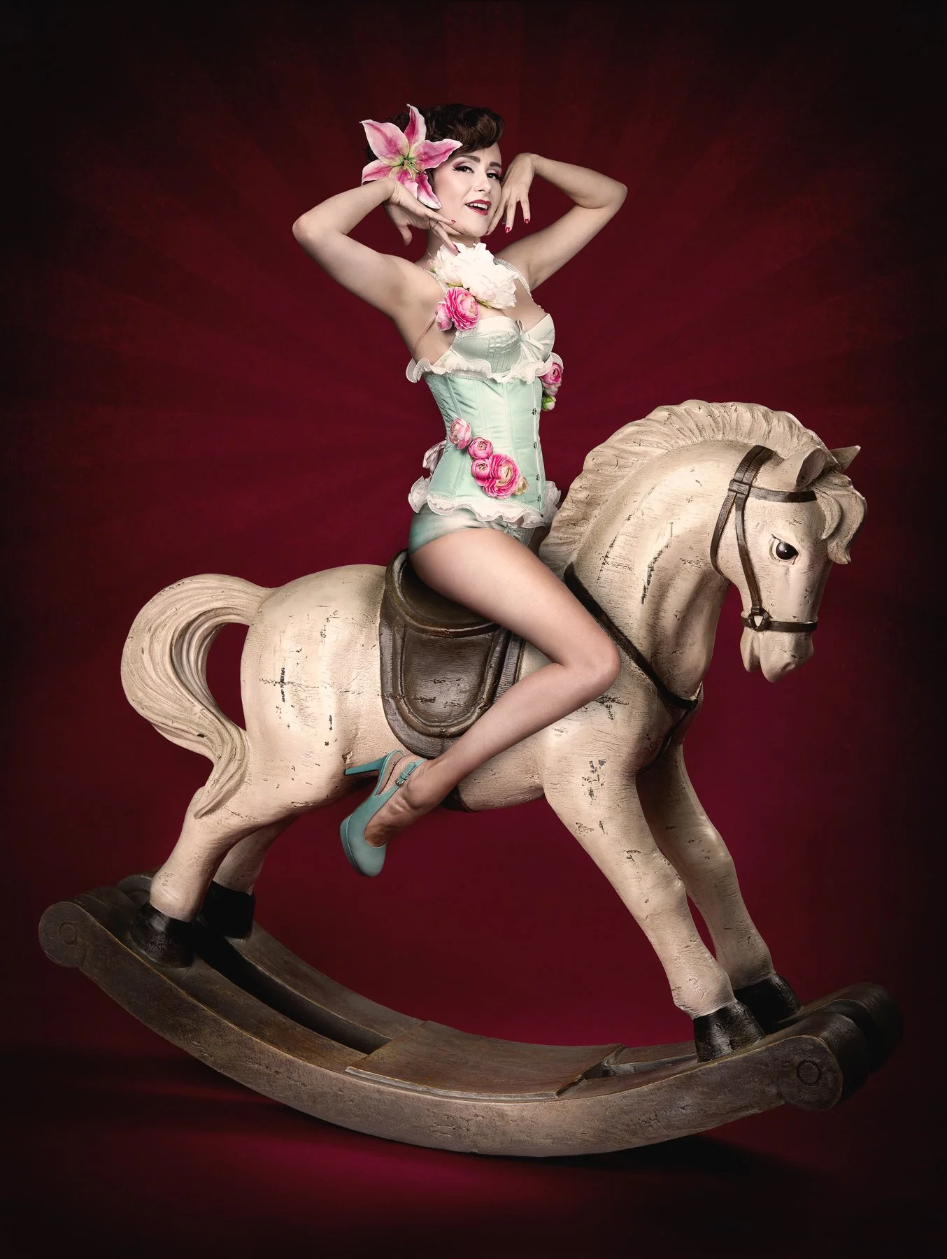 Une femme portant des vêtements en dentelle et des fleurs roses, assise sur un cheval à bascule en bois dans un studio avec un fond rouge foncé.
