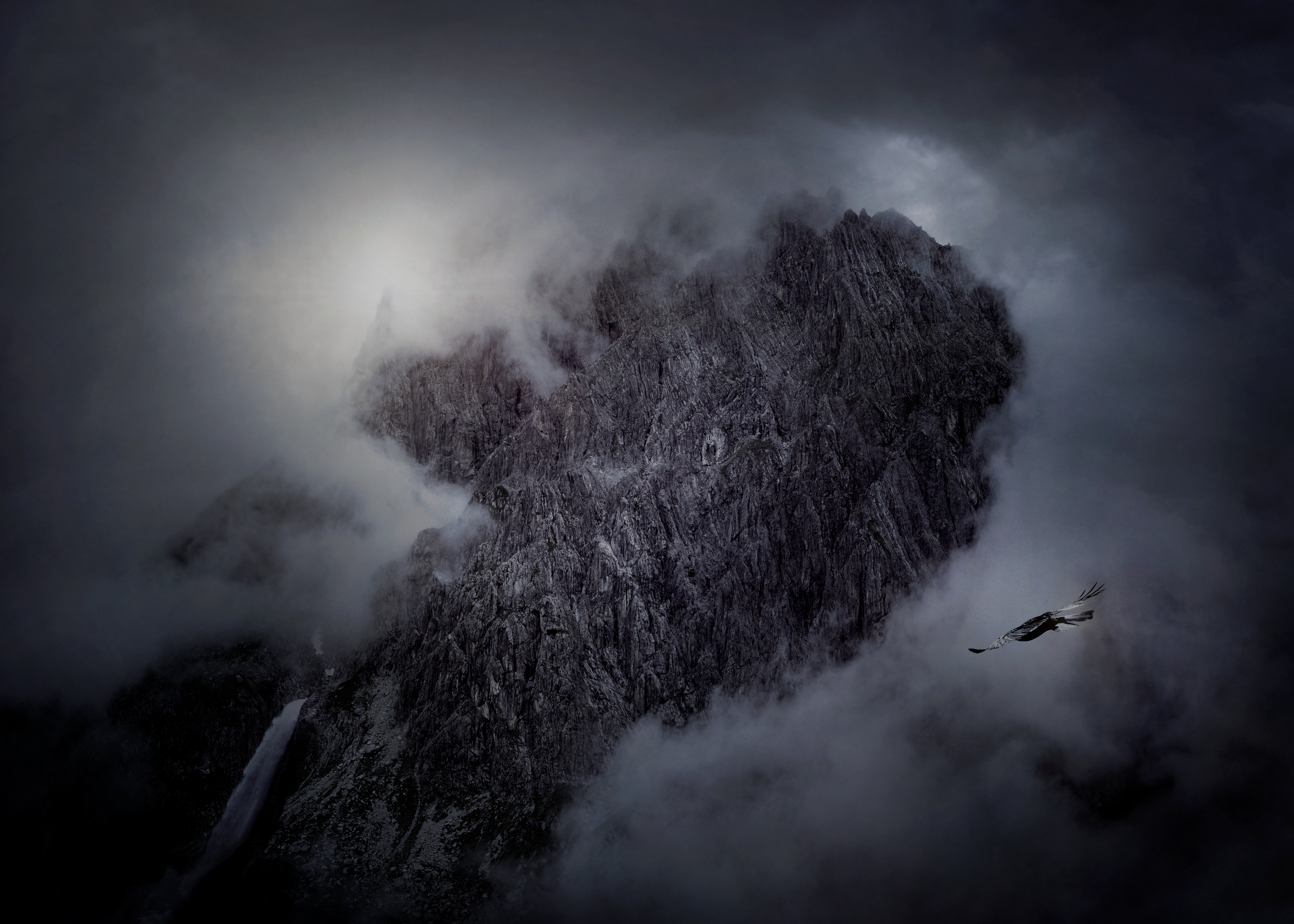 Une montagne escarpée entourée de nuages et de brume, avec une cascade au bas de la montagne. Il y a également un oiseau volant devant la scène.