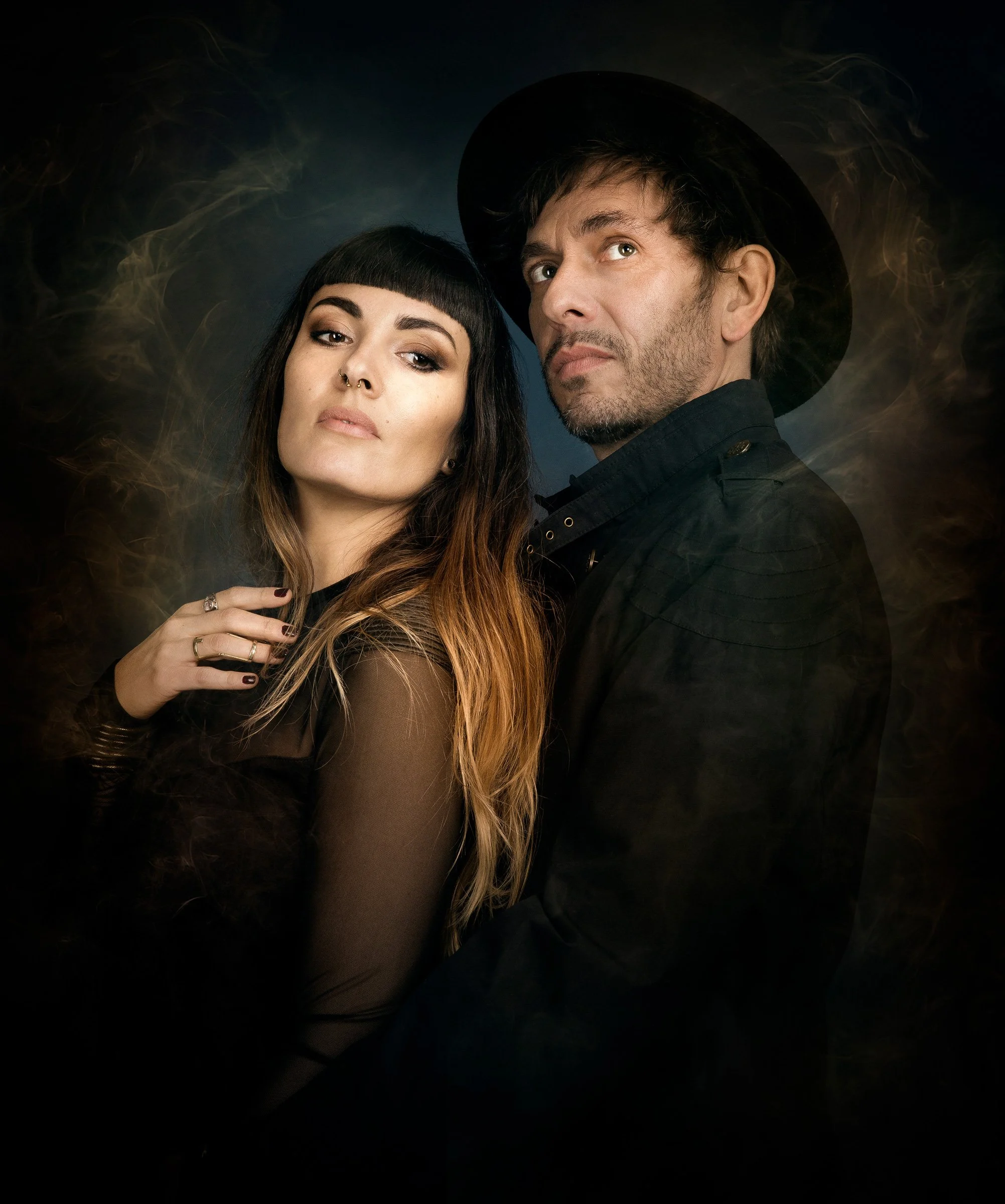 un homme et une femme posant côte à côte, la femme a des cheveux longs ombrés et un maquillage sombre, tandis que l'homme porte un chapeau noir et un manteau noir, fond sombre avec des volutes de fumée