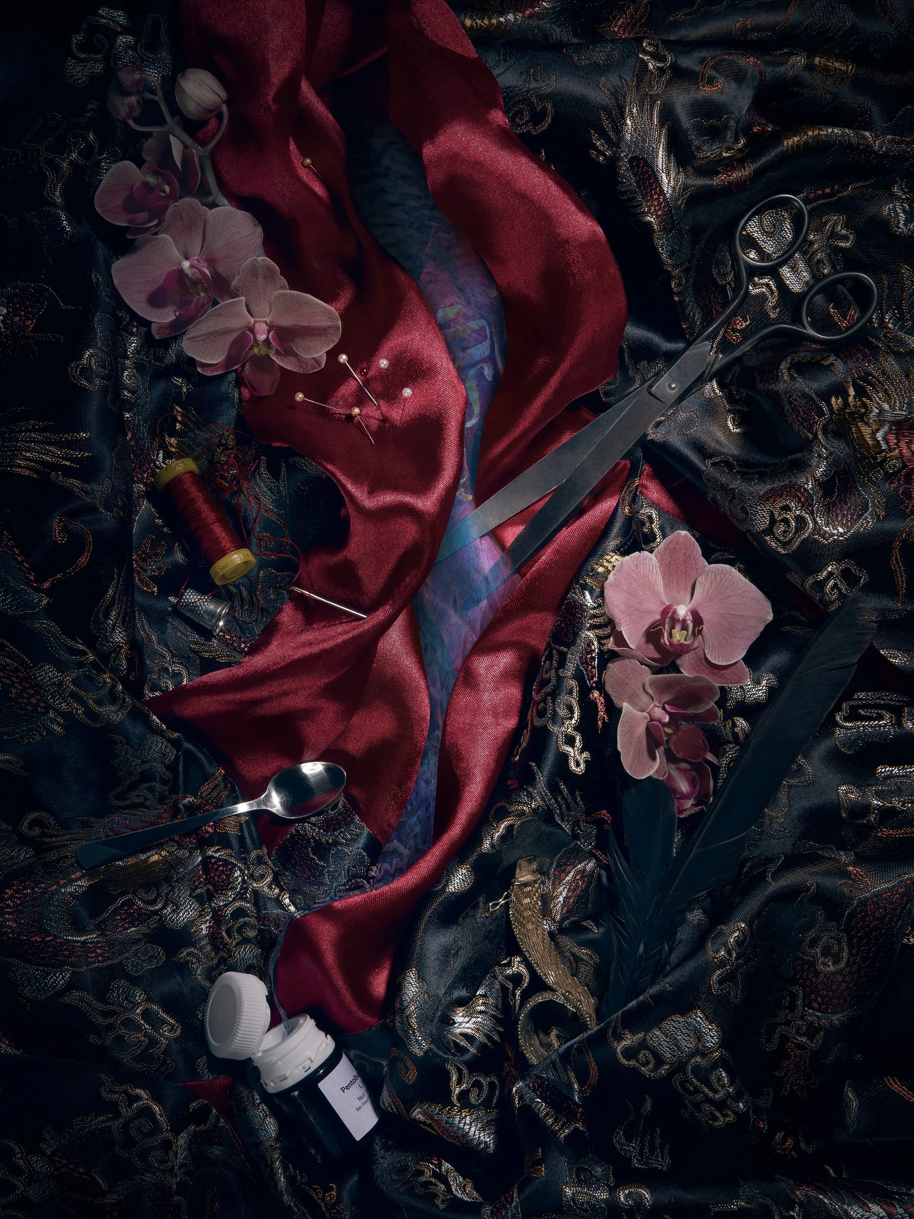 Une étoffe noire brodée de fils dorés avec une bordure en satin rouge, entourée de fleurs d'orchidée roses, des ciseaux, une bobine de fil rouge, une bouteille de solution désinfectante et d'autres éléments de couture.