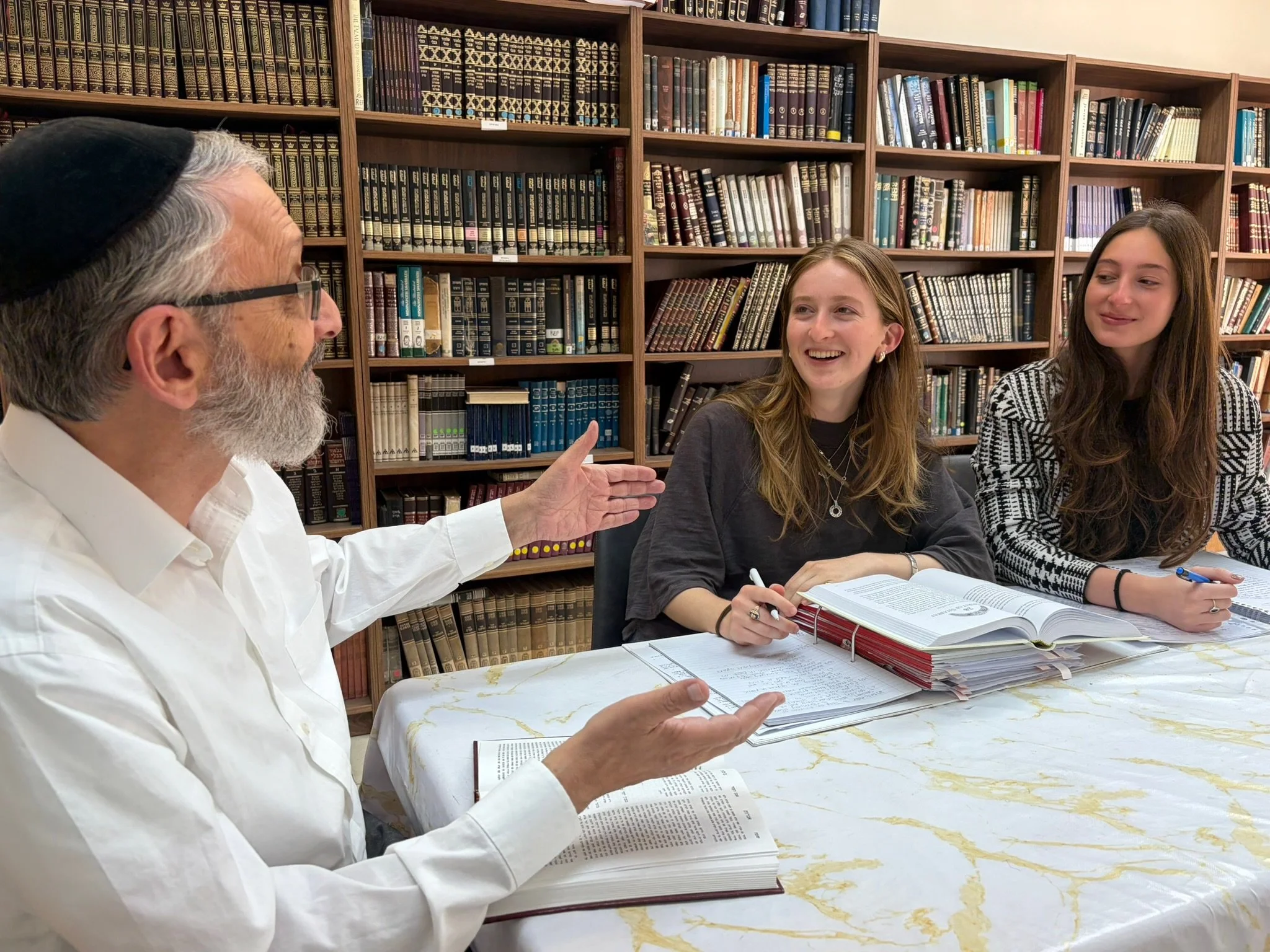 A man with a gray beard, glasses, and a black cap speaking to two young women sitting at a table in a library. The women are smiling and taking notes, with bookshelves filled with numerous books in the background.