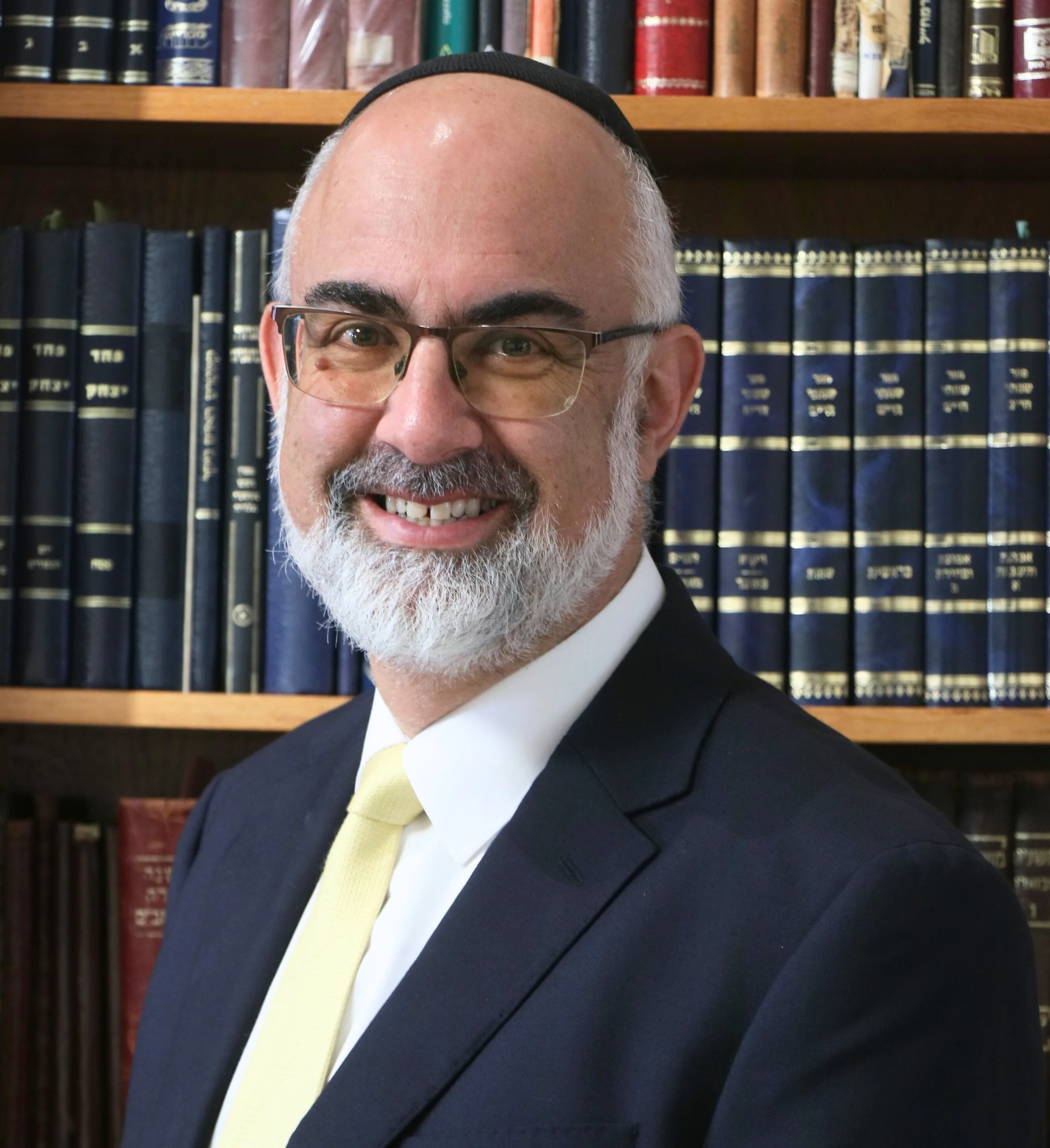 Smiling man with glasses and a beard, dressed in a dark suit and yellow tie, standing in front of a bookshelf filled with legal books.