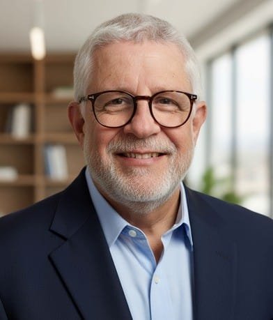 A smiling elderly man with glasses and gray hair, wearing a dark blazer and light blue shirt, in an office environment.