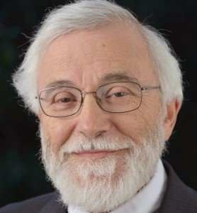 Close-up portrait of an elderly man with white hair, glasses, and a beard smiling against a dark background.