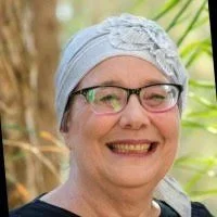 Smiling woman wearing glasses and a light blue headscarf, outdoors with green foliage in the background.