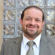 A man wearing a gray suit with a white shirt and a light gray tie, smiling in front of a glass block wall.