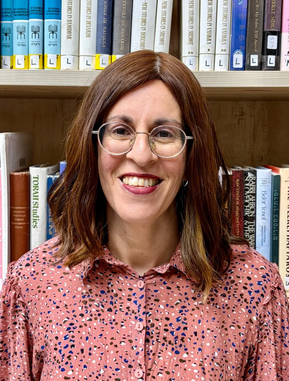 A woman with shoulder-length brown hair, wearing glasses and a pink patterned blouse, smiling in front of a bookshelf filled with books.