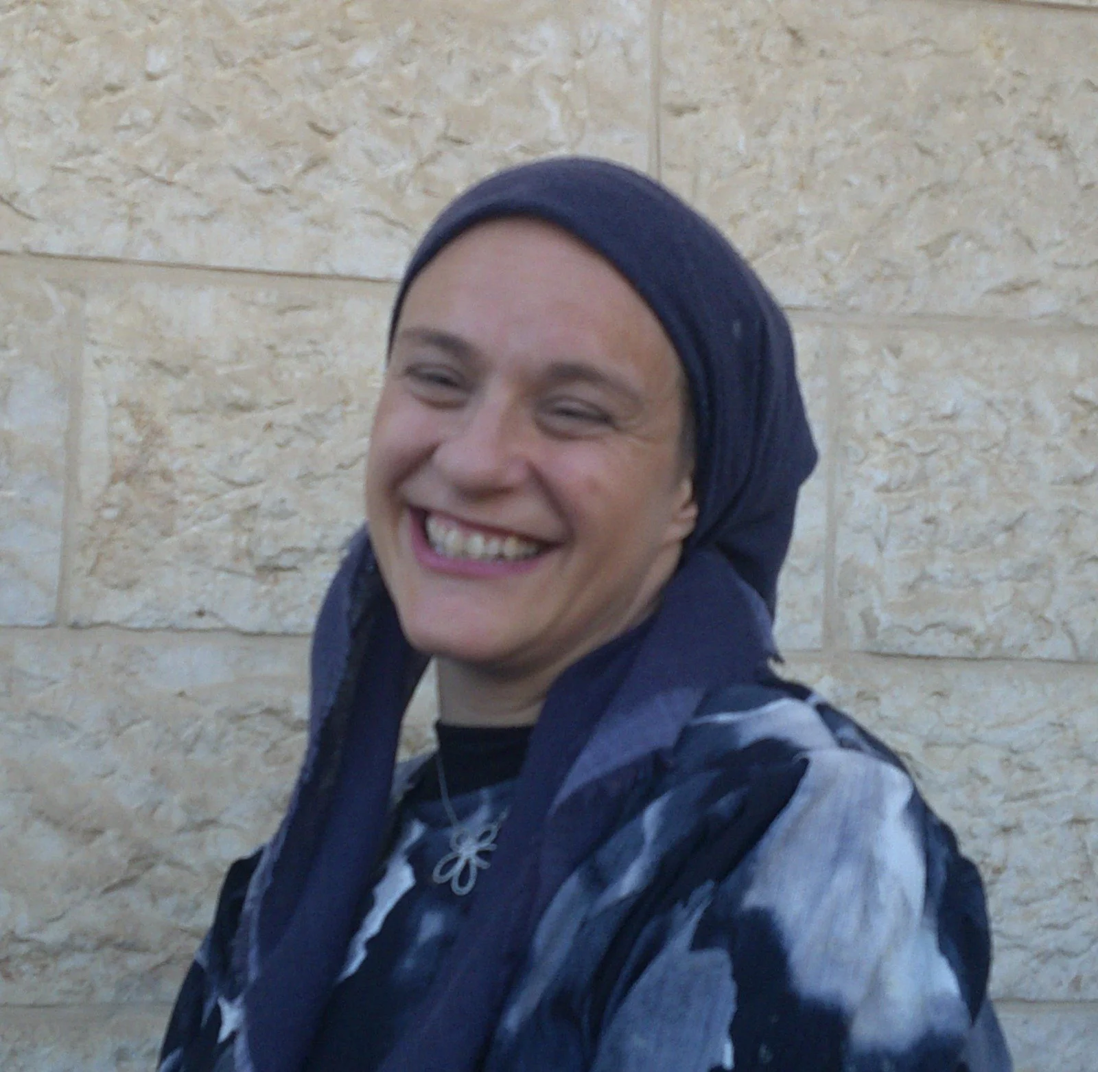 A smiling woman with a blue headscarf and a patterned jacket standing in front of a beige tiled wall.
