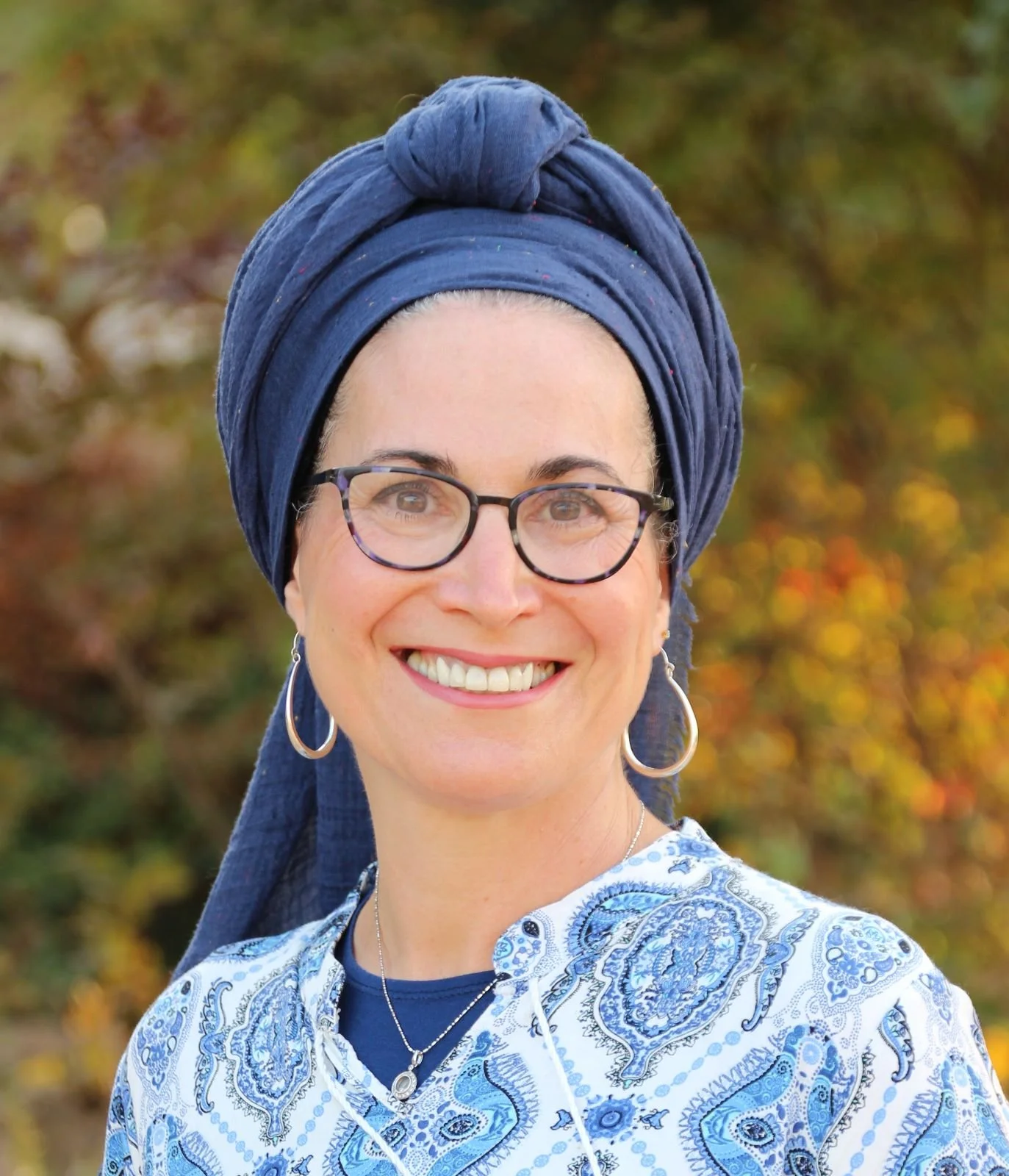 A smiling woman wearing glasses, a blue head wrap, and a blue and white paisley patterned blouse. She is outdoors with autumn trees in the background.