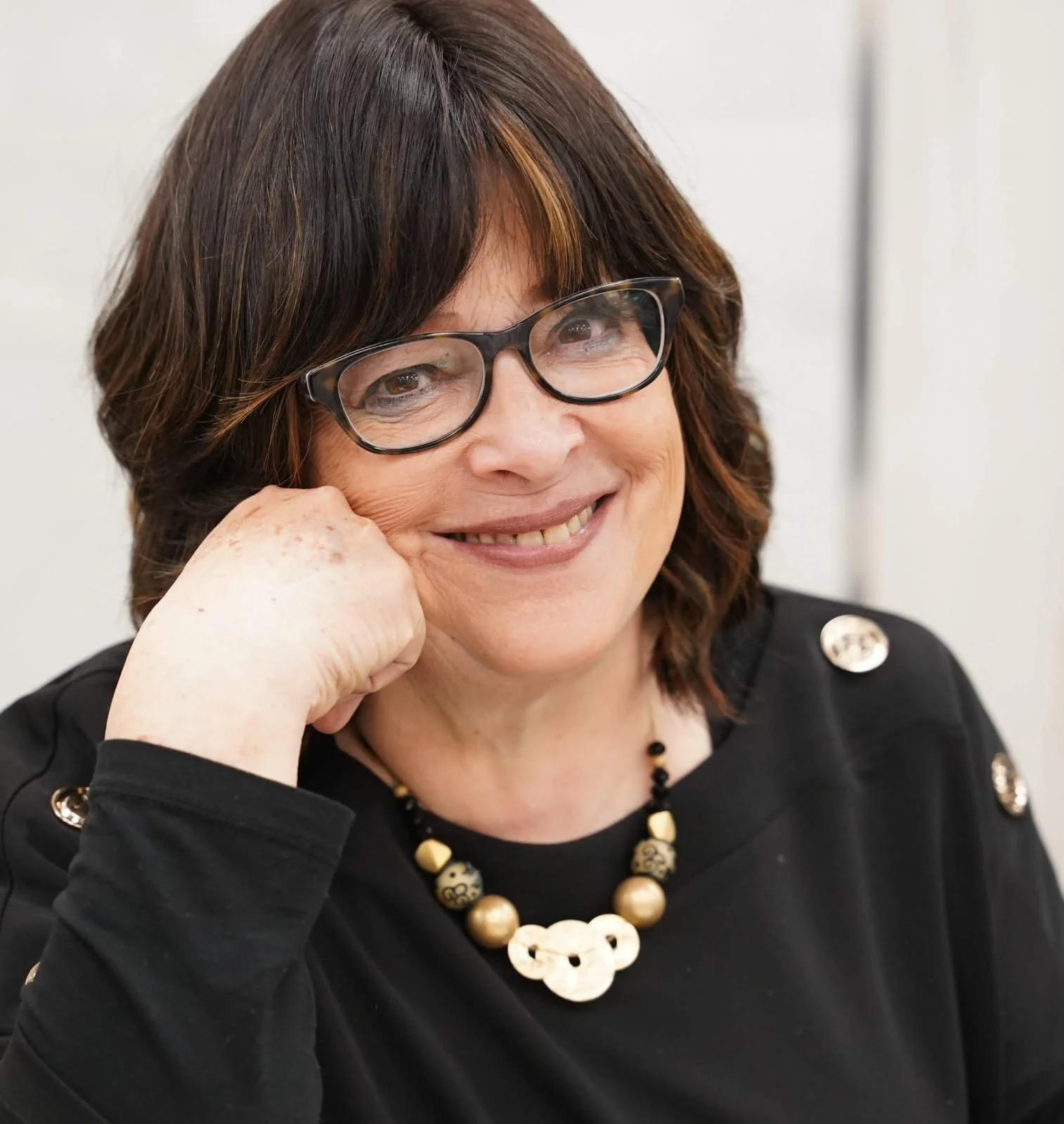 A woman with brown hair wearing glasses, smiling, resting her chin on her hand, dressed in a black top with gold accents and a beaded necklace.