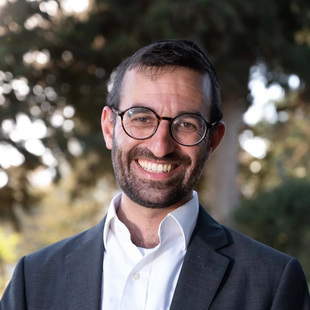 A smiling man in a suit and glasses standing outdoors with trees in the background.