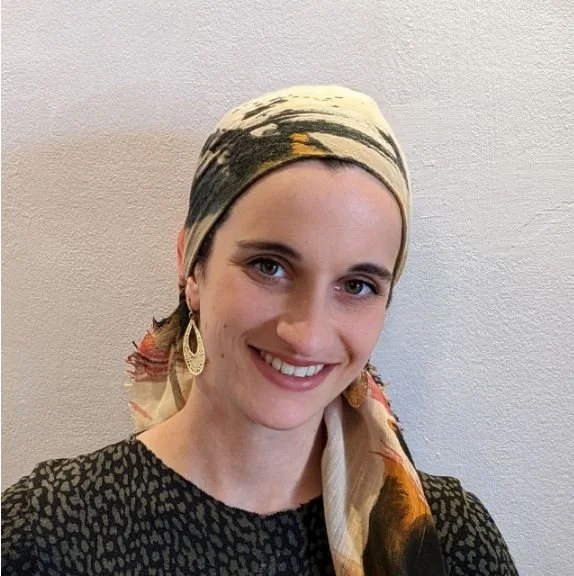 A woman smiling, wearing a patterned headscarf and large earrings, against a plain wall.