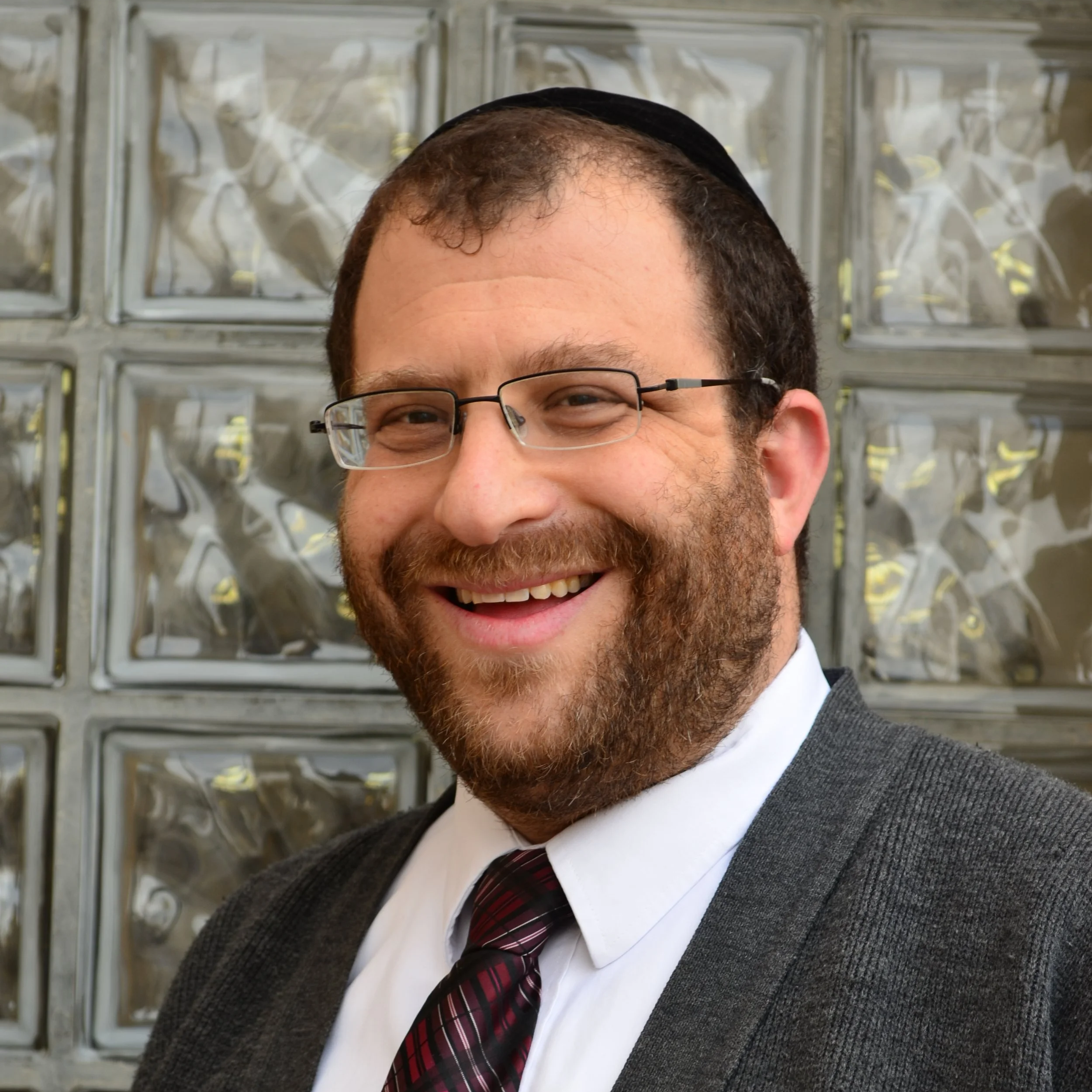 Close-up of a man wearing glasses, a white shirt, a dark red and black tartan tie, and a gray plaid blazer, smiling in front of glass blocks.