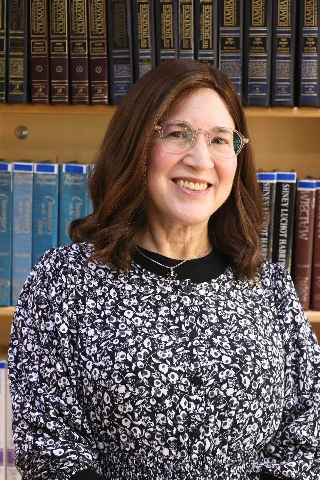 A woman with brown hair and glasses smiling in front of shelves filled with books.