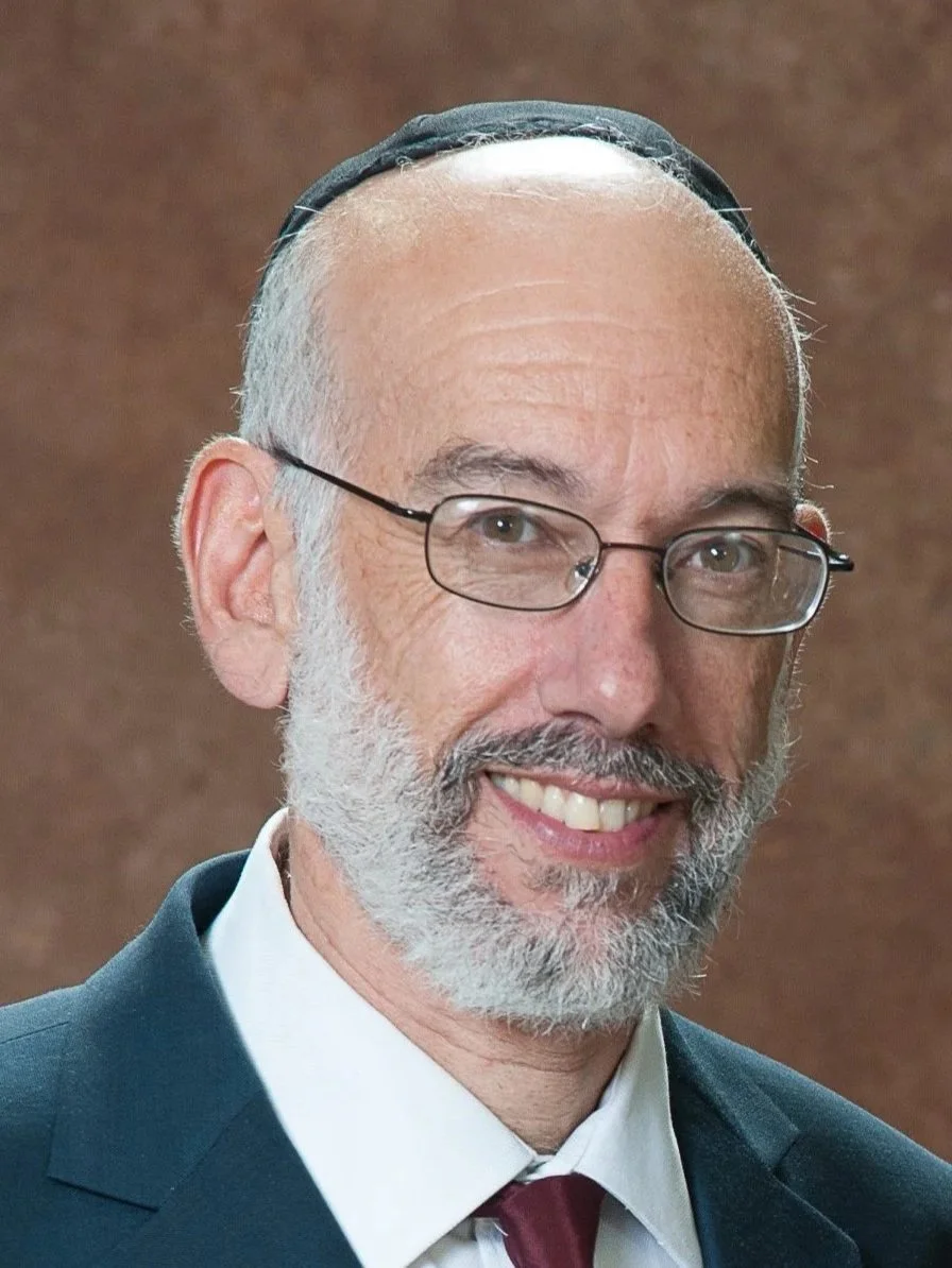 A smiling man with glasses, gray beard, wearing a dark suit, white shirt, maroon tie, and a yarmulke.