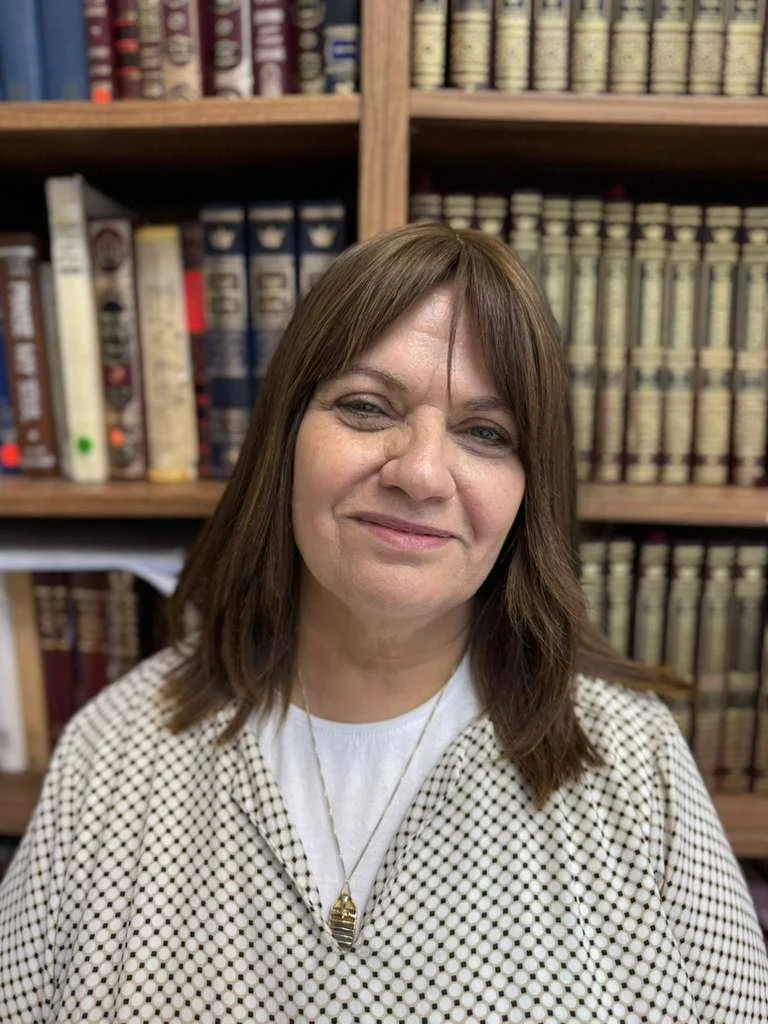 A woman with shoulder-length brown hair, wearing a white shirt with black polka dots and layered necklaces, standing in front of a bookshelf filled with legal books.