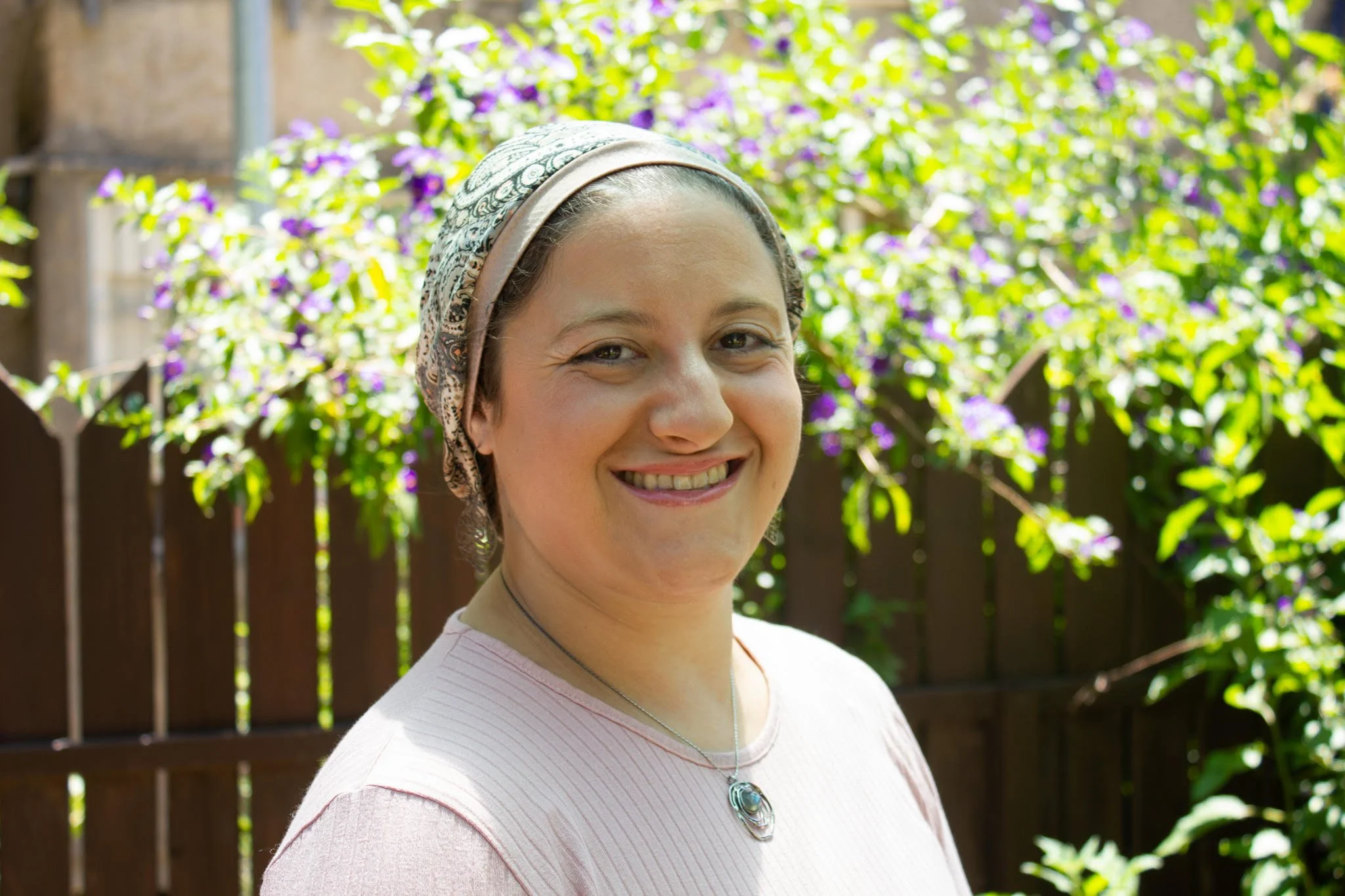 Smiling woman with head scarf standing outdoors in front of a garden fence and blooming purple flowers.