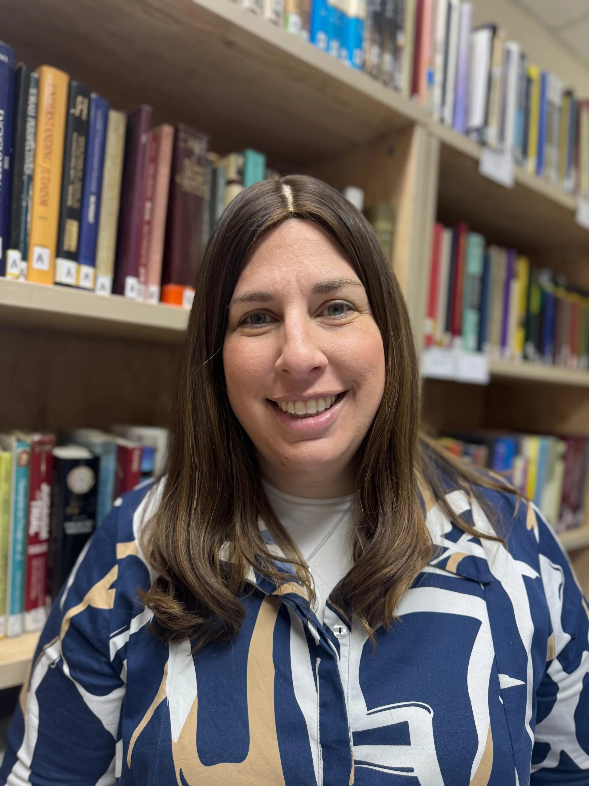 A smiling woman with long brown hair standing in front of a bookshelf in a library.