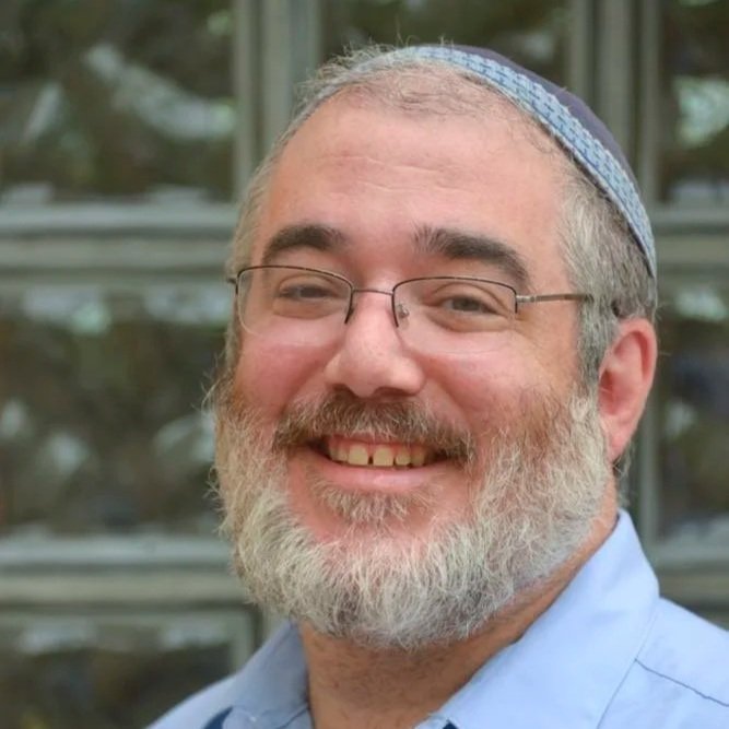 A smiling man with glasses, a grey and white beard, wearing a light blue shirt and a blue yarmulke, standing outdoors with blurred greenery and windows in the background.