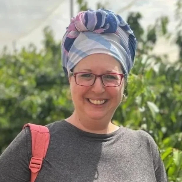 A woman smiling outdoors, wearing glasses, a gray shirt, and a colorful head wrap, with a pink backpack strap visible on her shoulder, and green foliage in the background.