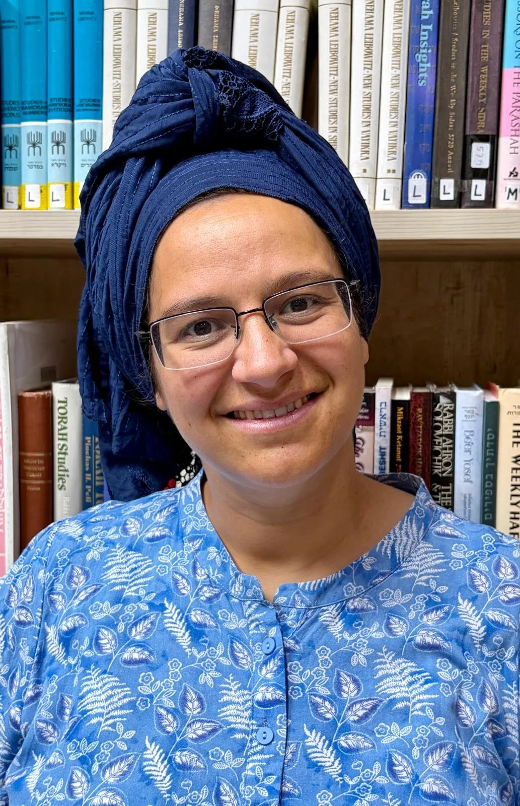 A person with glasses smiling, wearing a blue patterned shirt and a large blue headwrap, standing in front of a bookshelf.