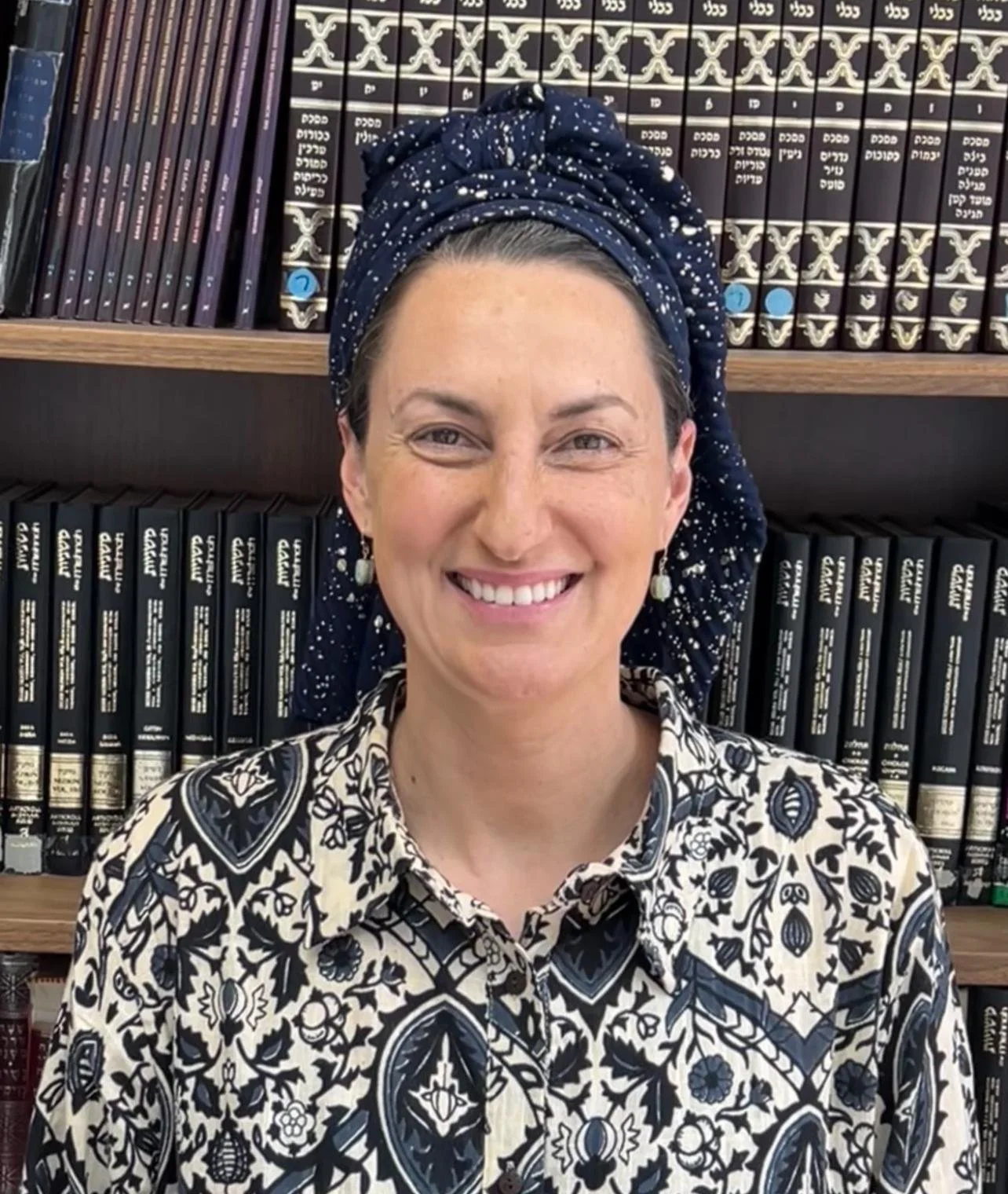 A woman with a patterned shirt and a headscarf smiling in front of a bookshelf filled with books.