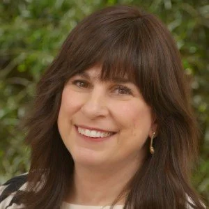 A woman with shoulder-length brown hair and bangs smiling outdoors with greenery in the background.