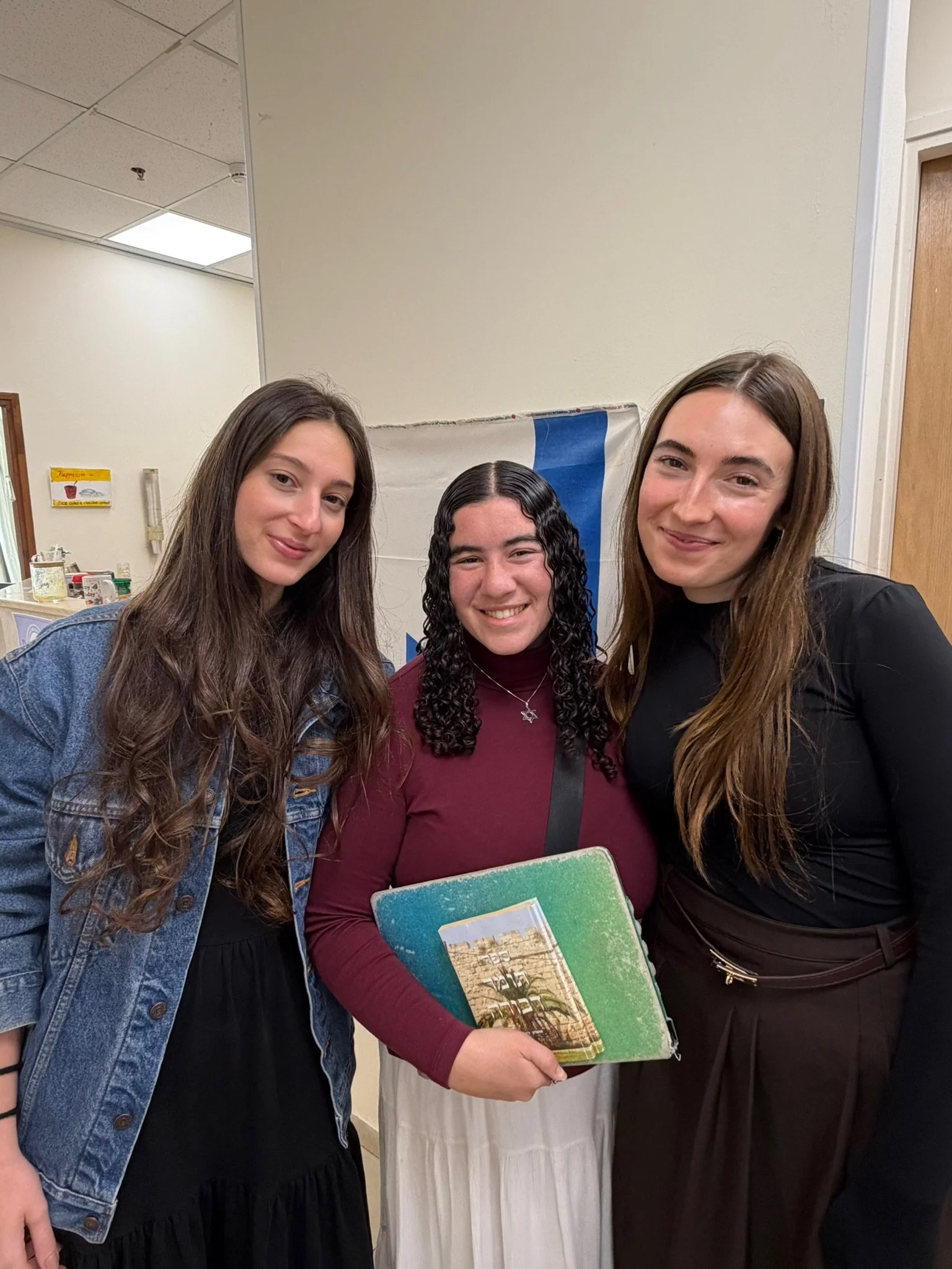 Three women standing close together, smiling, in an indoor setting. The woman in the middle is holding a colorful book or folder.