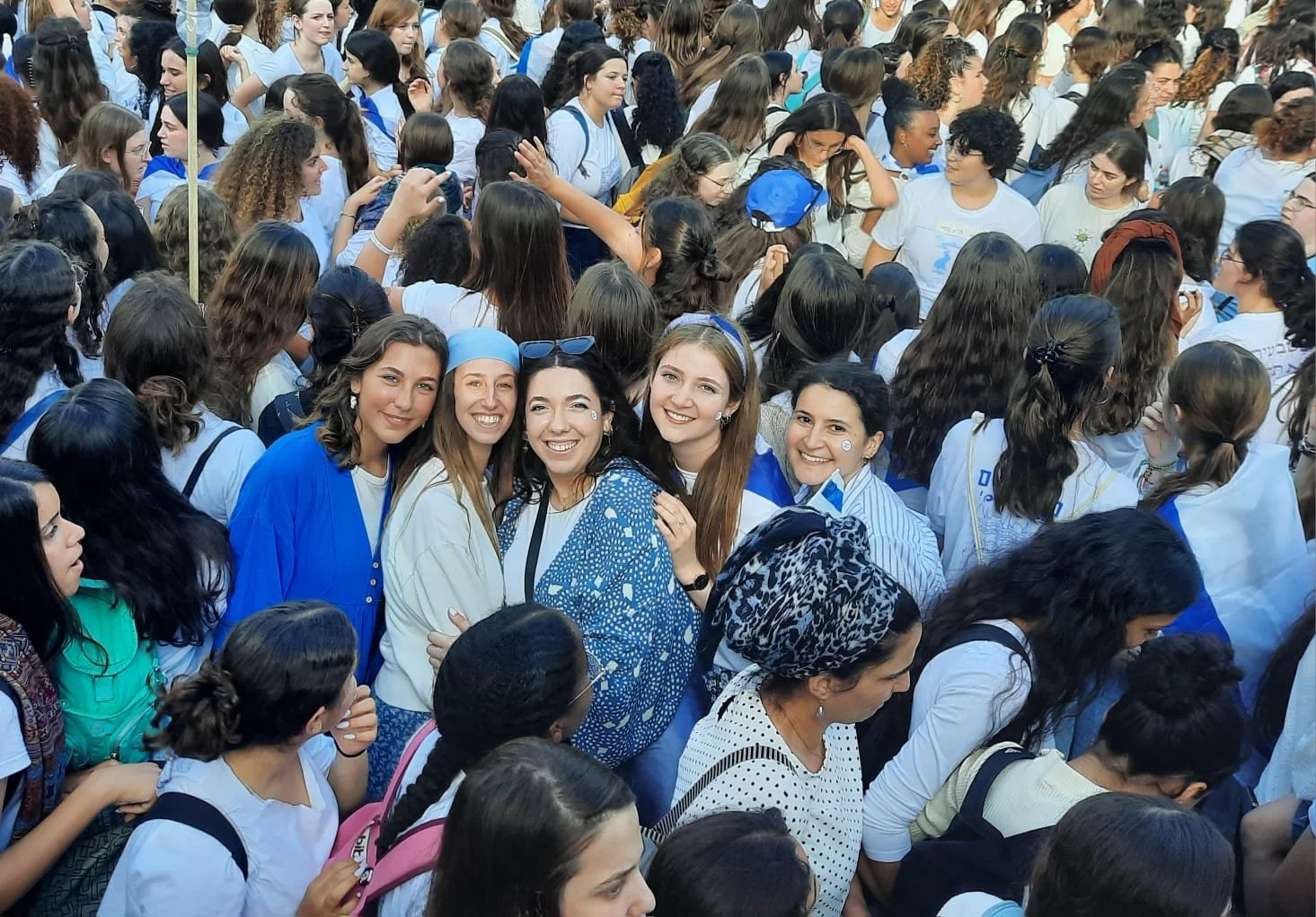 A large group of women and girls gathered outdoors, dressed mostly in white and blue, some wearing head coverings or accessories, with a few smiling and posing for the camera in the foreground.