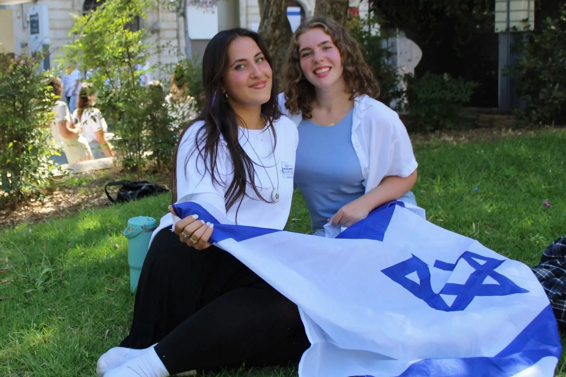 Two women sitting on grass outside, holding a flag with blue and white colors, smiling at the camera. In the background, additional groups of people are gathered near trees and a house.