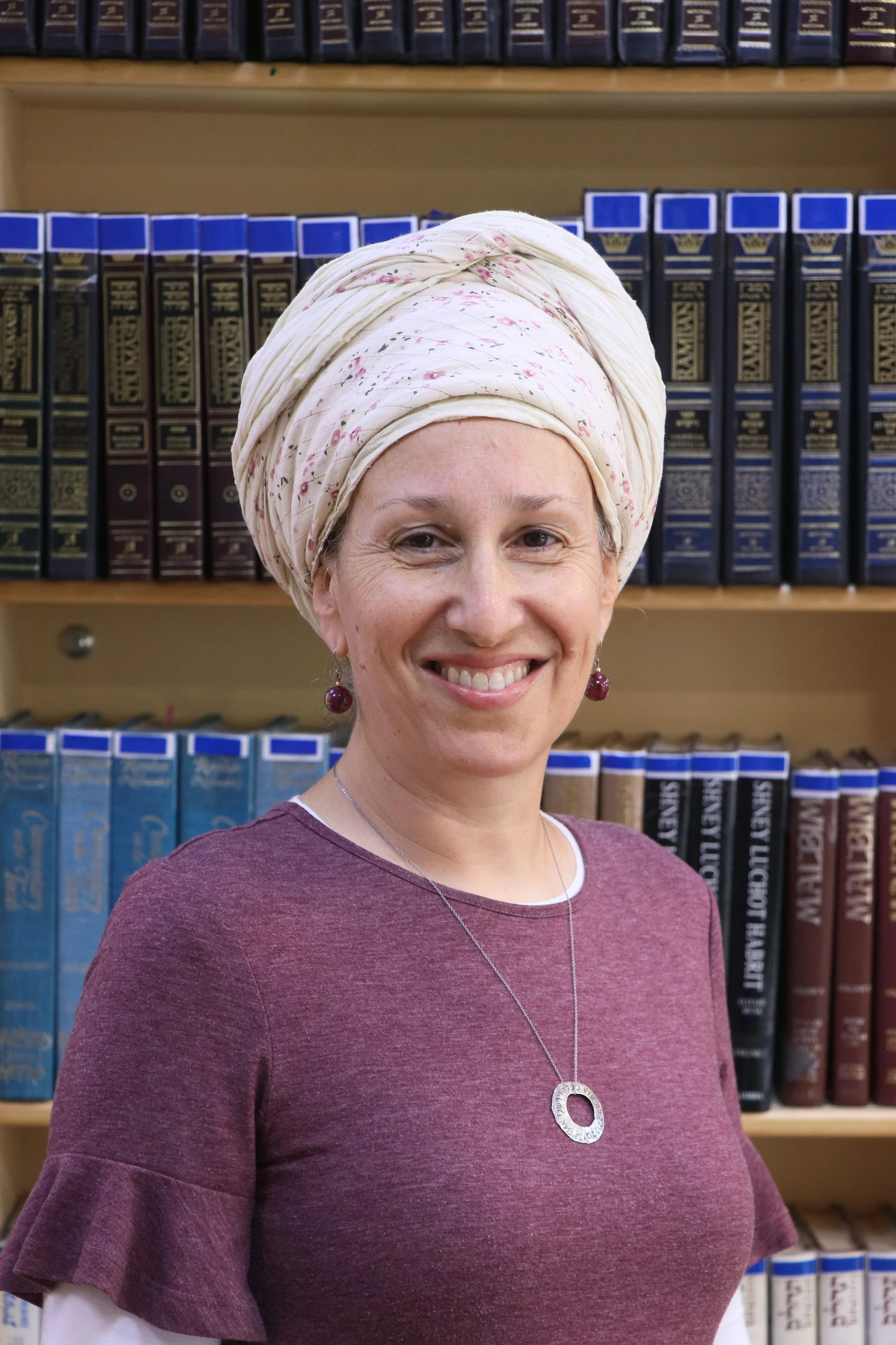 A smiling woman with a light complexion, wearing a maroon top with ruffled sleeves and a pendant necklace, standing in front of a bookshelf filled with law books.