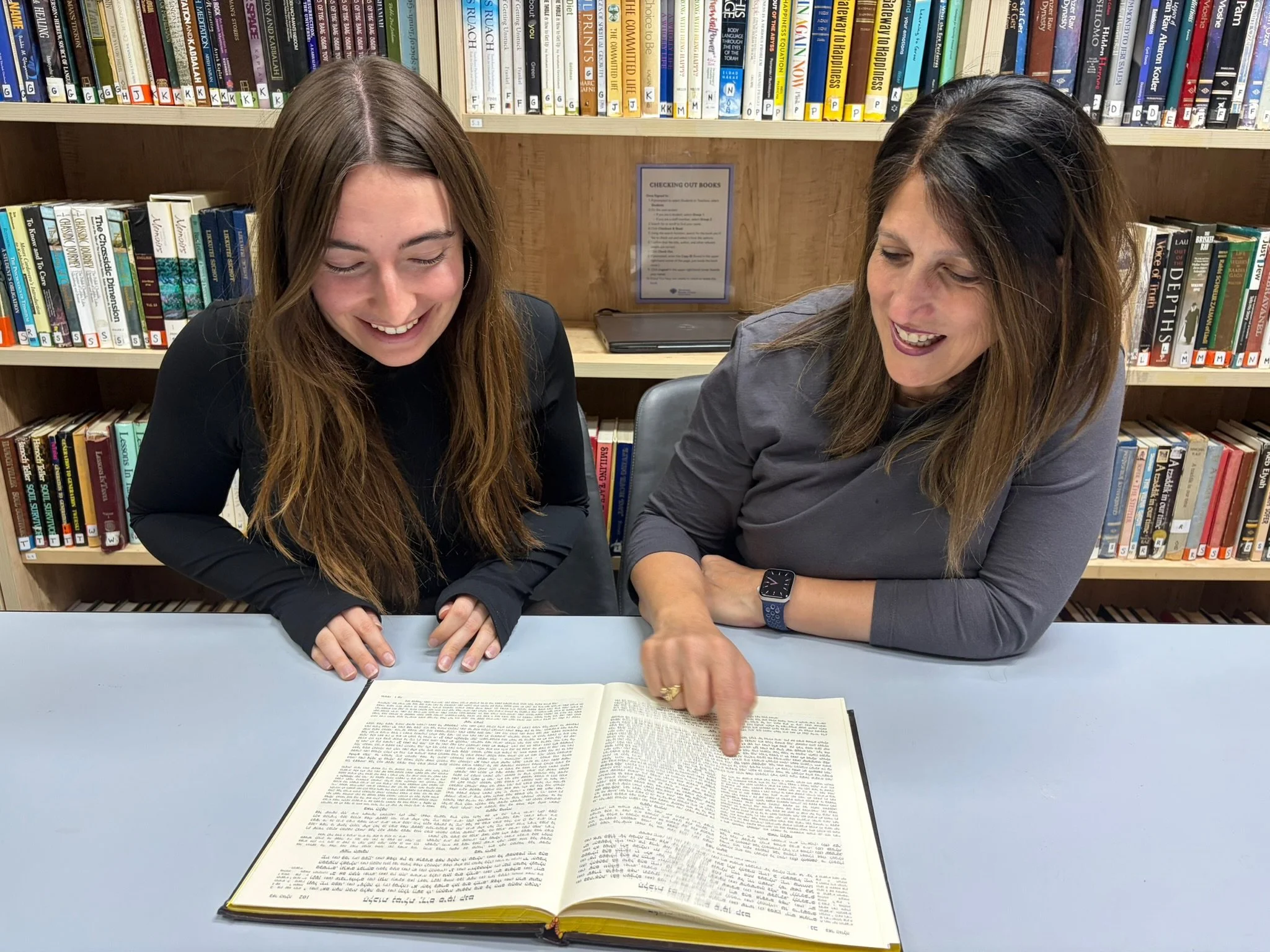 Two women sitting at a table in a library, looking at an open large book with text, smiling and pointing at the pages. bookshelf with many books is in the background.