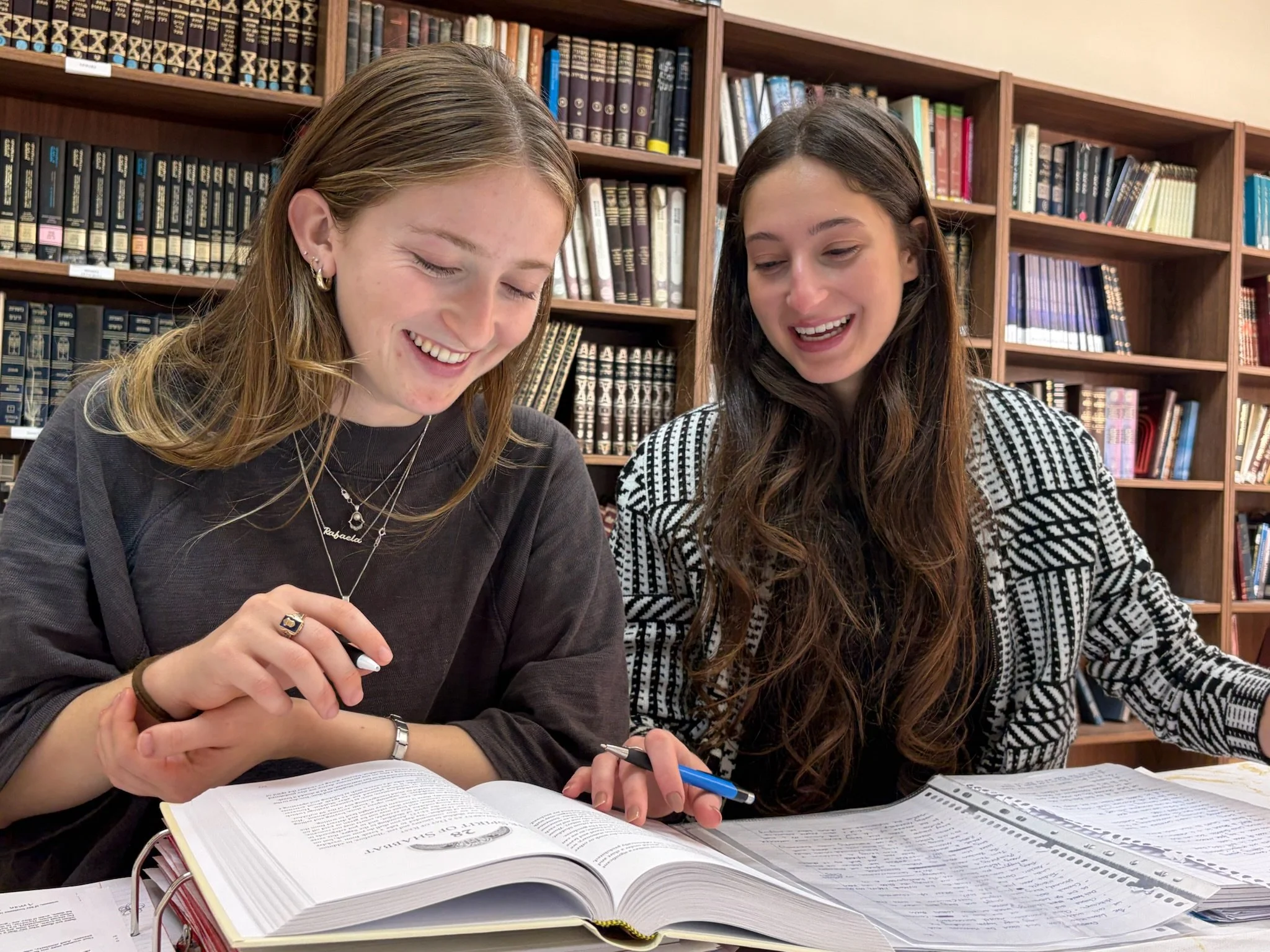 Two young women sit at a table in a library, laughing and studying together with open textbooks and notebooks.