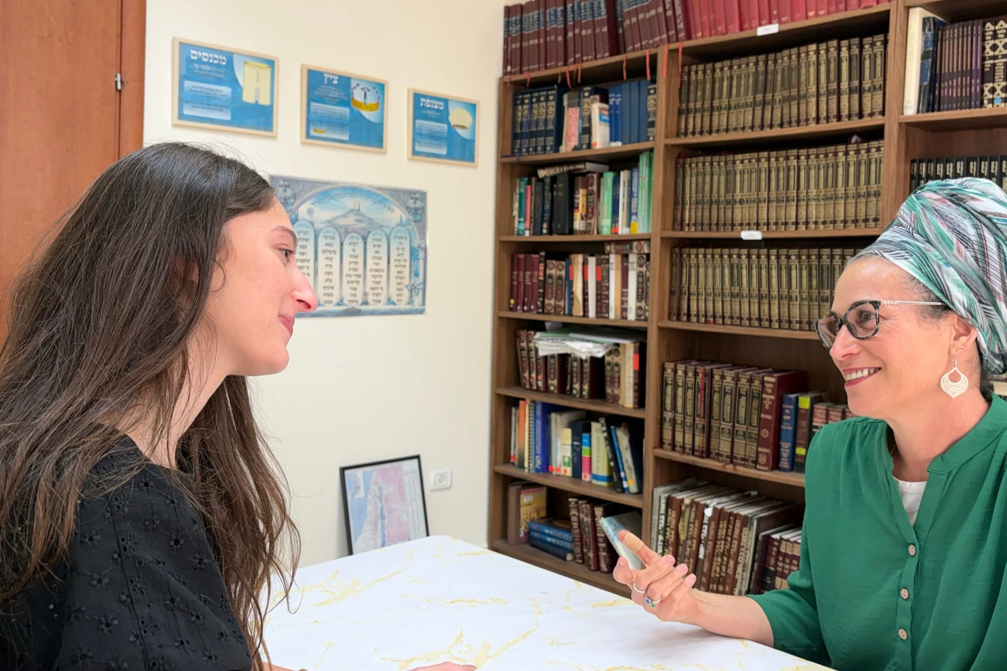 A woman with glasses and a headscarf smiling while talking to a young woman with long brown hair, in a room with bookshelves filled with books.