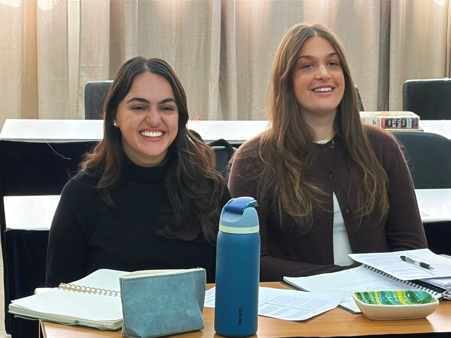 Two women sitting at a conference table, smiling. On the table are notebooks, a pen, papers, a water bottle, and a small bowl. A third woman is partially visible in the background. The setting appears to be a meeting or study session.
