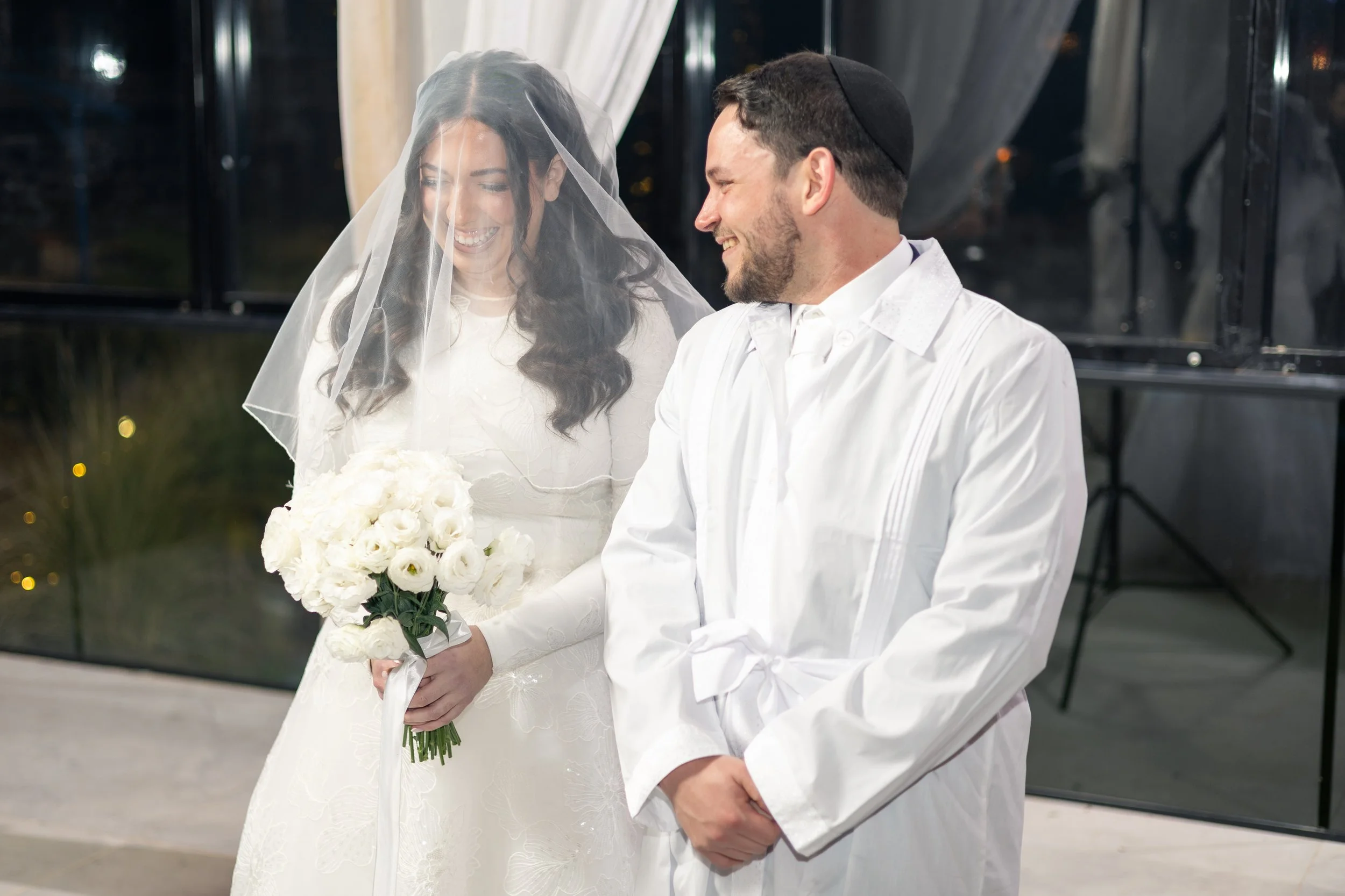 A bride and groom smiling during their wedding ceremony. The bride is holding a bouquet of white roses and wearing a veil, while the groom is wearing a white shirt and a kippah.
