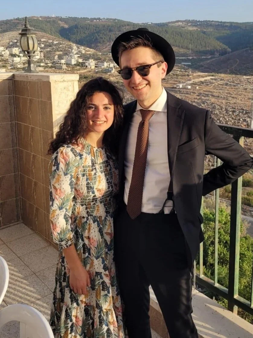 A smiling woman with wavy dark hair in a colorful floral dress standing next to a smiling man wearing sunglasses, a black hat, a black suit, white shirt, and brown tie, on a balcony overlooking a valley with hills and scattered buildings.